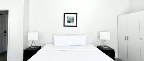 A minimalist hotel room with a large bed, two bedside tables with lamps, a wardrobe, and a framed picture on the wall.