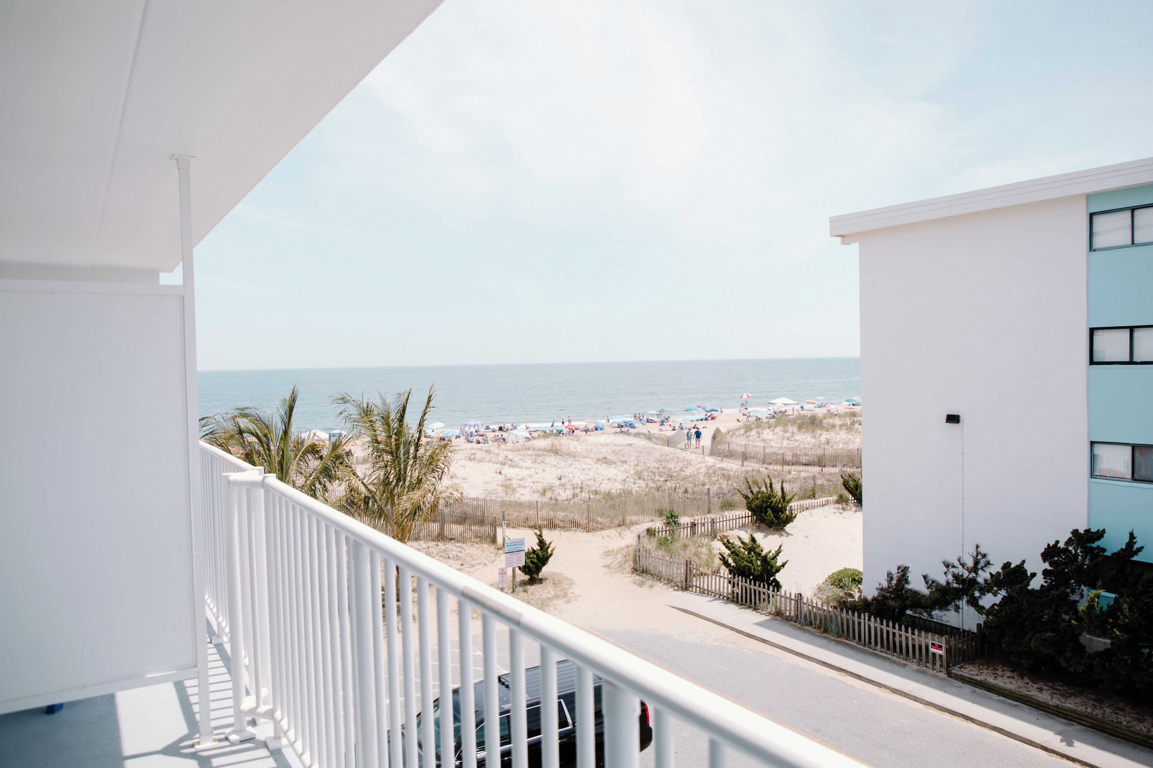 A balcony with a view of the beach, a fence, and a building with a light blue facade.