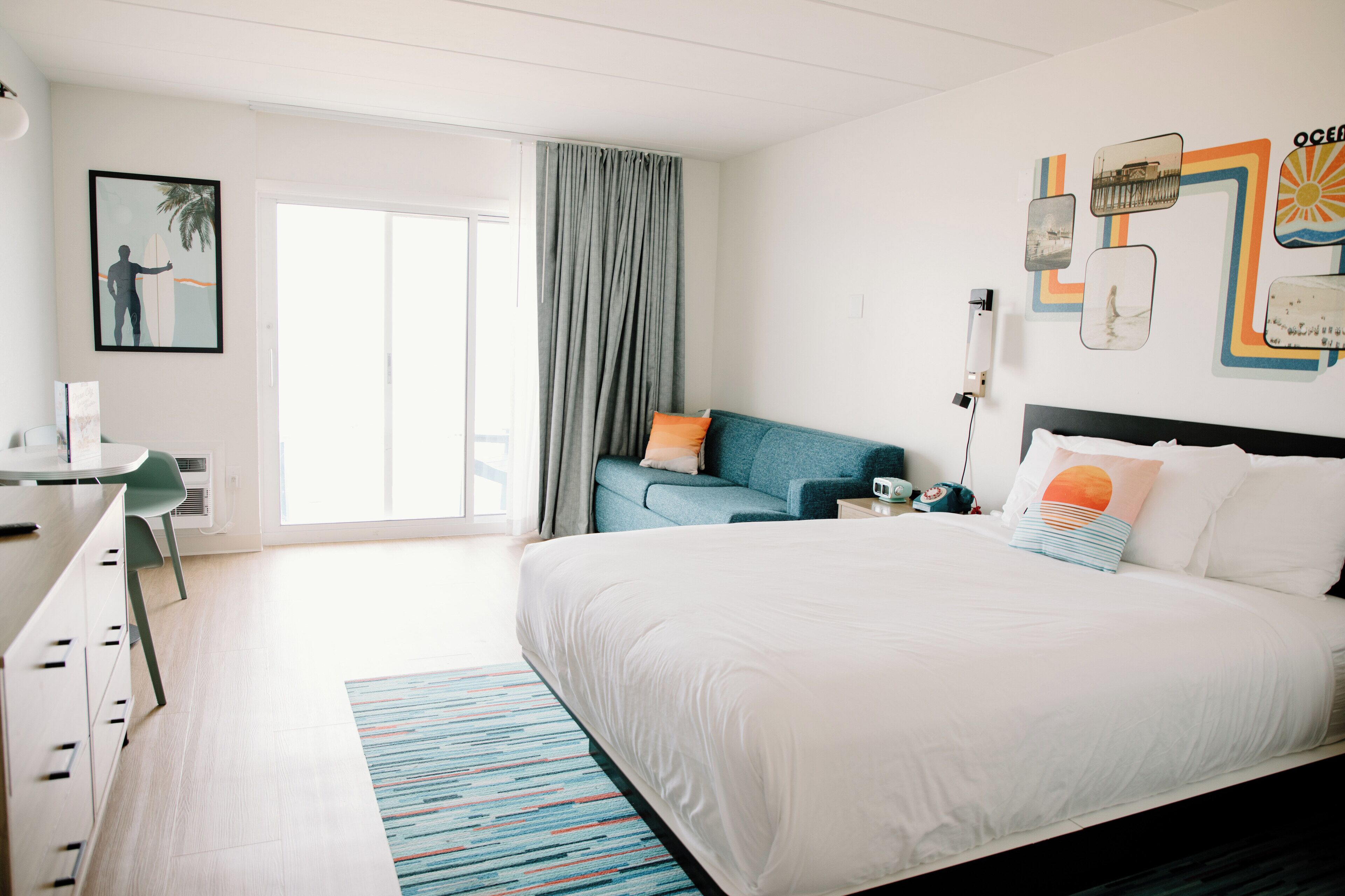 A modern hotel room with a large bed, a desk, and a chair. There is a window with curtains, a wall with artwork, and a door leading to another room.