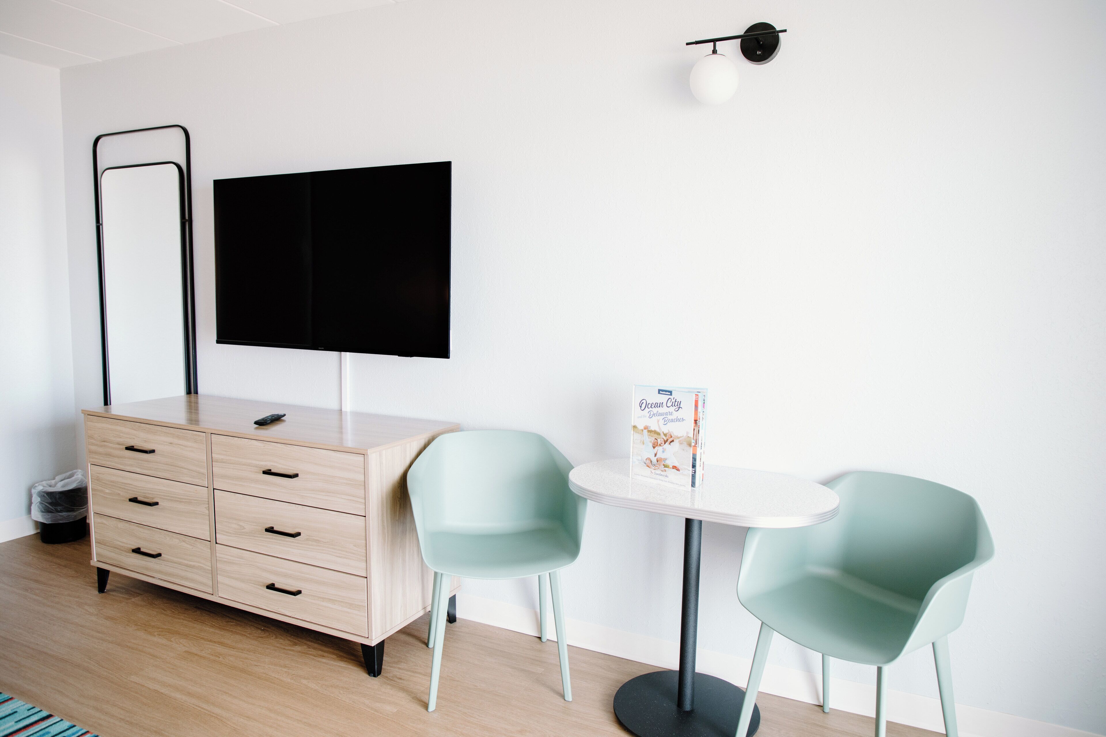 A modern room with a wooden dresser, a flat-screen TV, two light blue chairs, a small round table, and a trash bin.