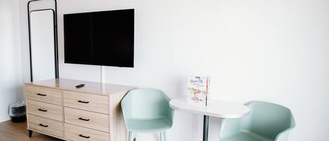 Une chambre moderne avec une commode en bois, un téléviseur à écran plat, deux chaises bleu clair, une petite table ronde et une poubelle.
