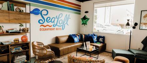 Un hall d’hôtel moderne avec une grande enseigne murale indiquant « SeaLoft Oceanfront Hotel », des meubles en osier, un canapé en cuir et une vue sur la rue à travers une grande fenêtre.