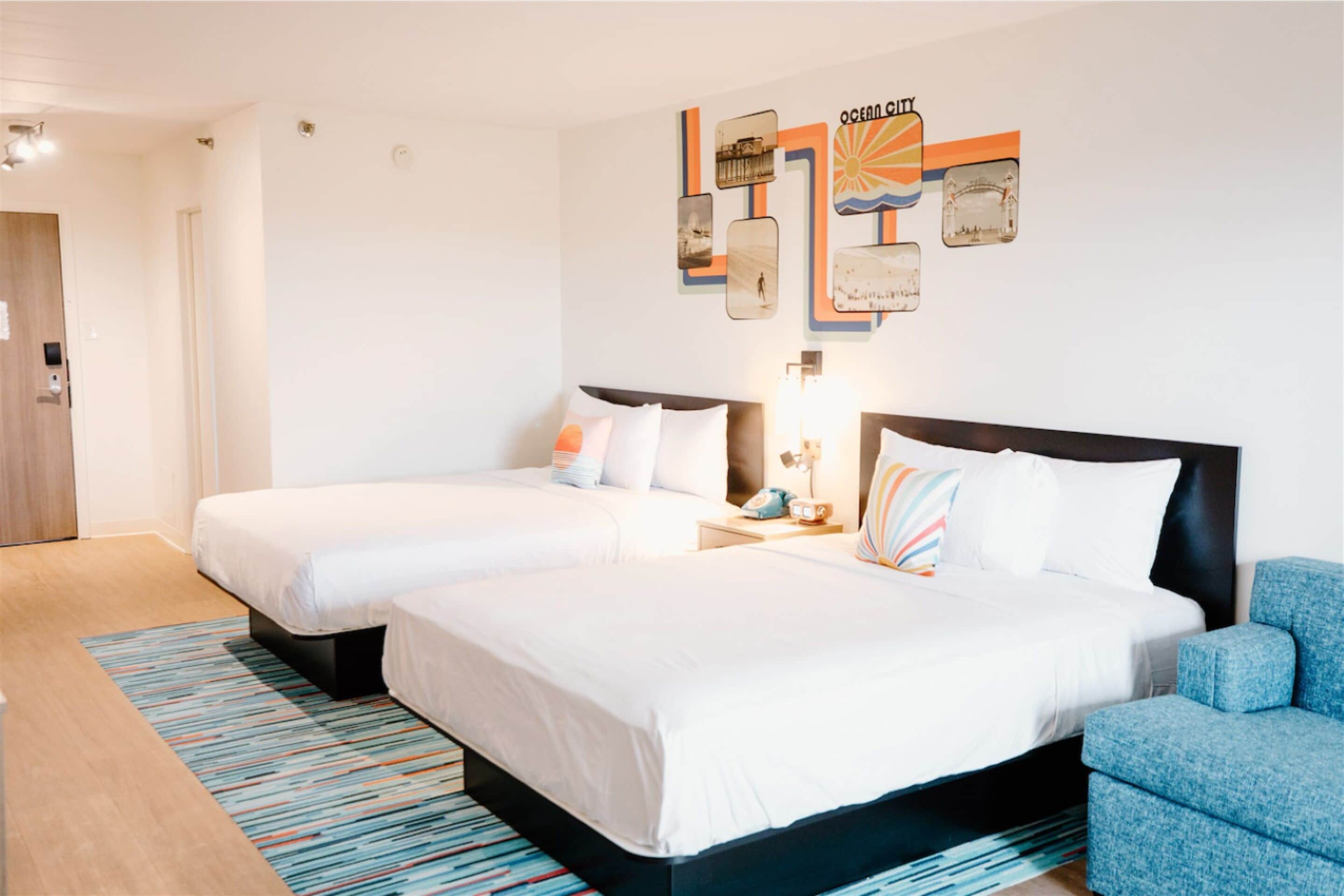 Habitación de hotel con dos camas, un sillón azul, un tapete a rayas de colores y una obra de arte en la pared.