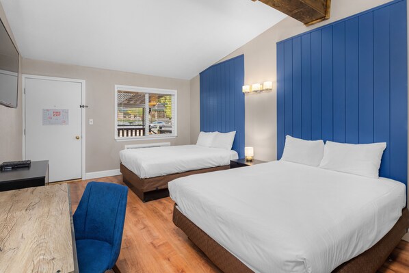 A hotel room with two beds, a wooden table, a blue chair, and a window with a view of the outside. - The Inn at Boatworks (Tahoe City)