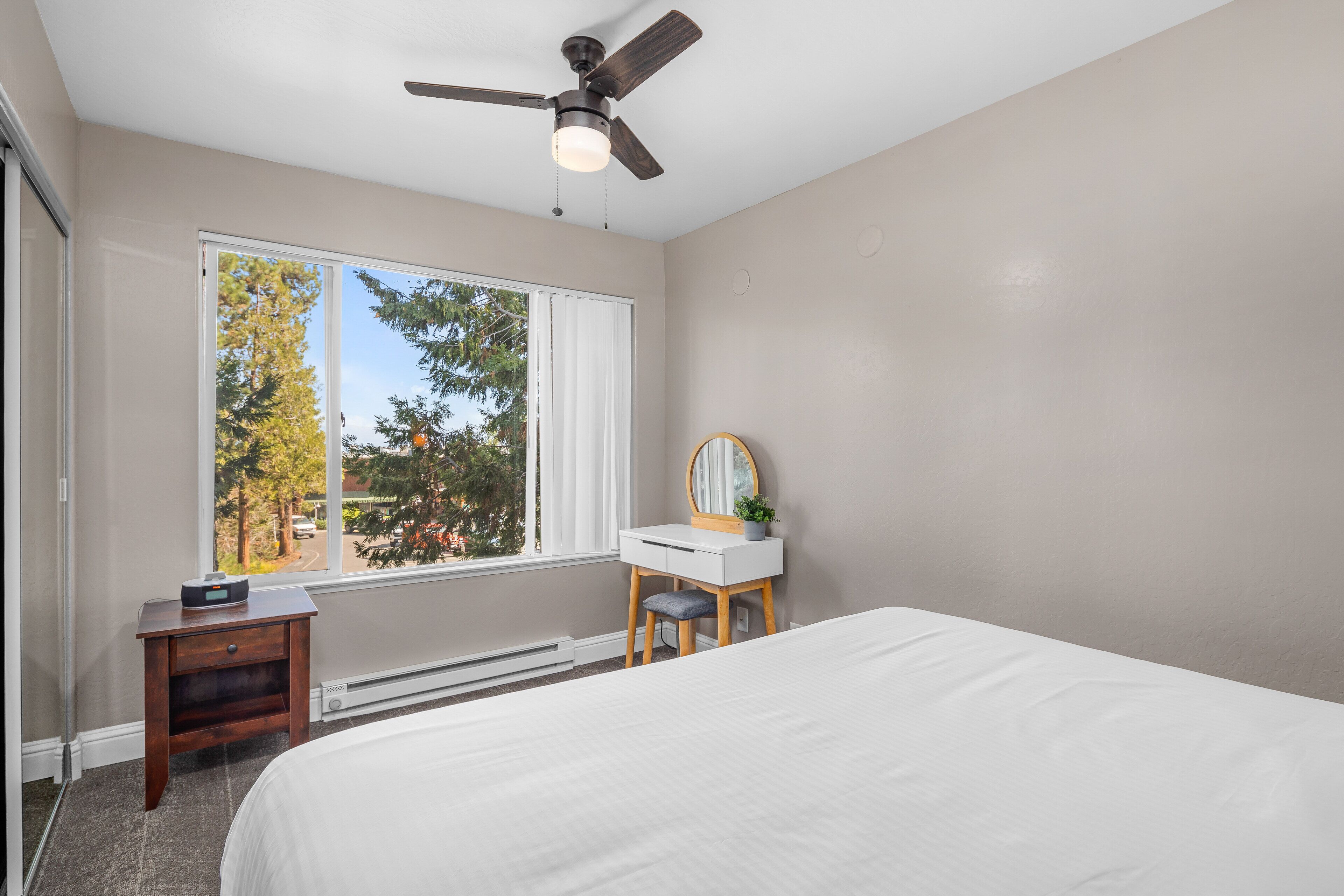 Un dormitorio con cama, escritorio, silla, ventilador de techo y ventana con vista a árboles y una carretera.