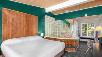 A bedroom with a large bed, a wooden headboard, a bathroom with a glass shower, and a view of the outdoors.