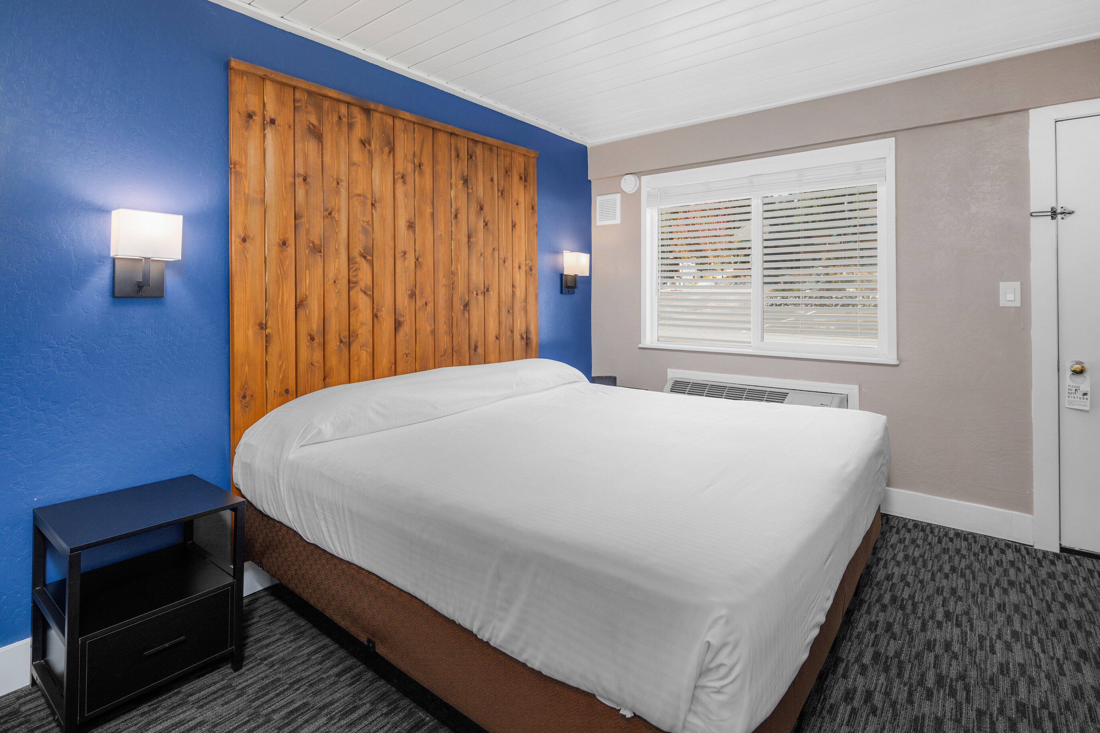 A bedroom with a wooden headboard, a bed with white bedding, a nightstand with a drawer, and a window with blinds.