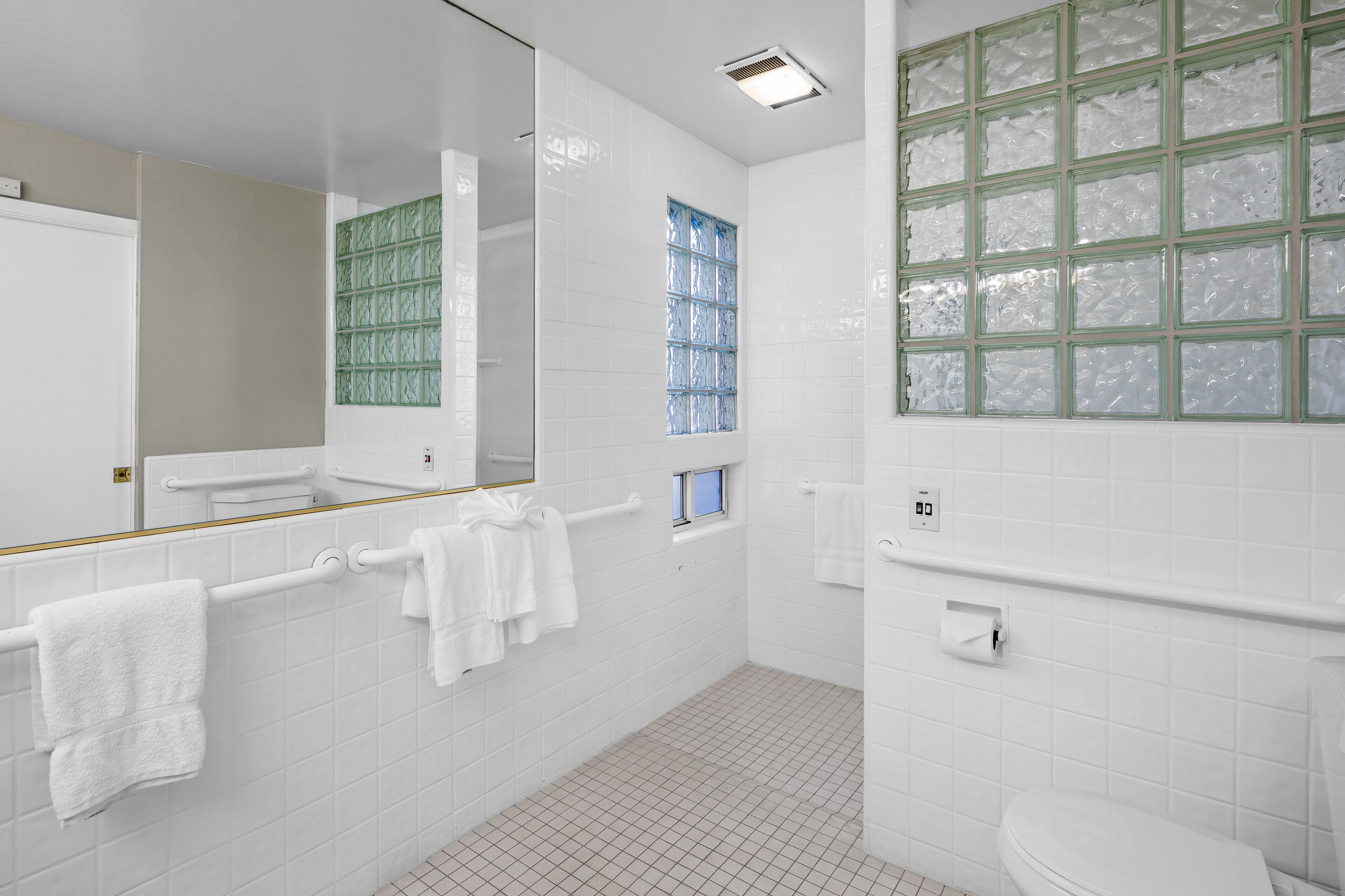 A modern bathroom with white tiles, a large mirror, a window with green frames, and a toilet with a white tank.