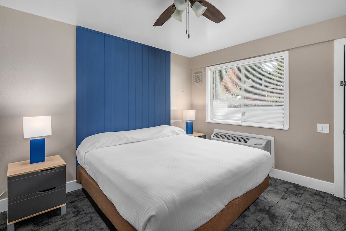 Une chambre à coucher comprenant un lit, deux tables de chevet avec des lampes, un ventilateur de plafond, une fenêtre avec des stores et un radiateur fixé au mur.