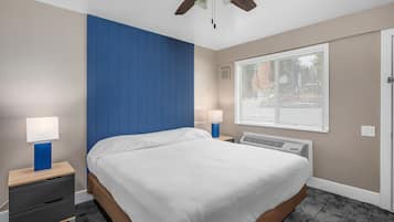 Quarto com cama, duas mesas de cabeceira com luminárias, ventoinha no teto, janela com persianas e aquecedor fixado na parede.