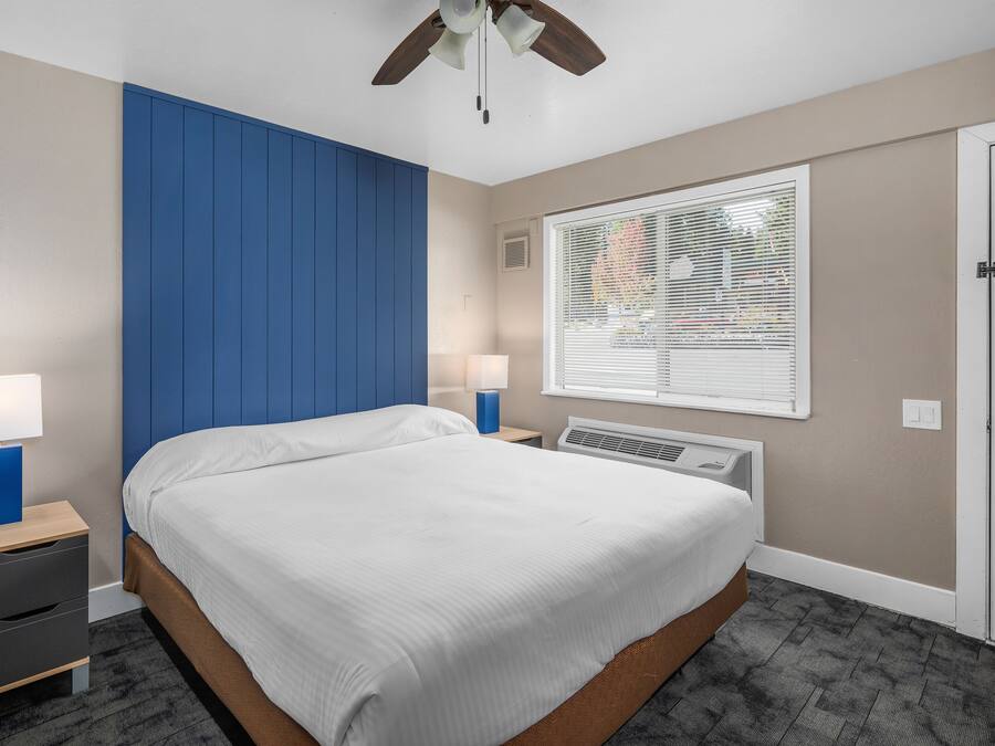 Quarto com cama, duas mesas de cabeceira com luminárias, ventoinha no teto, janela com persianas e aquecedor fixado na parede.