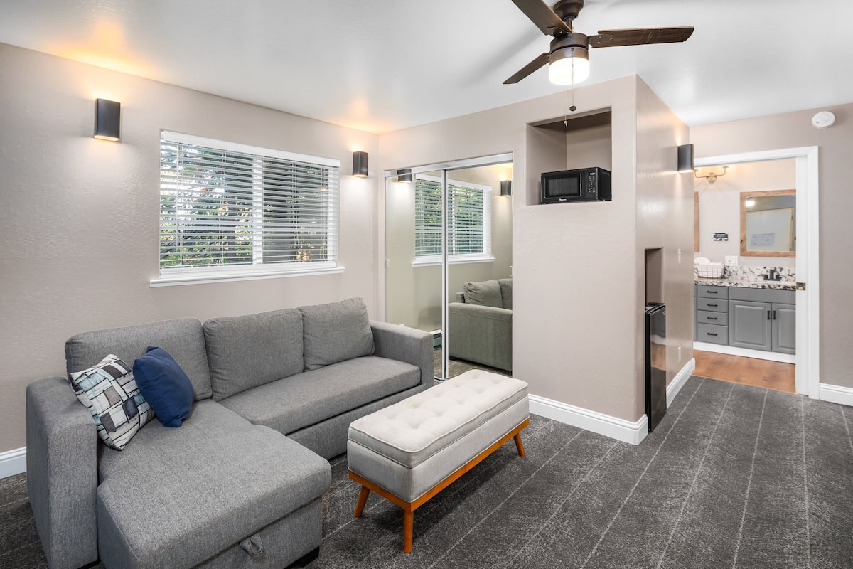 Un salon moderne avec un canapé d’angle gris, un pouf assorti et un ventilateur de plafond.