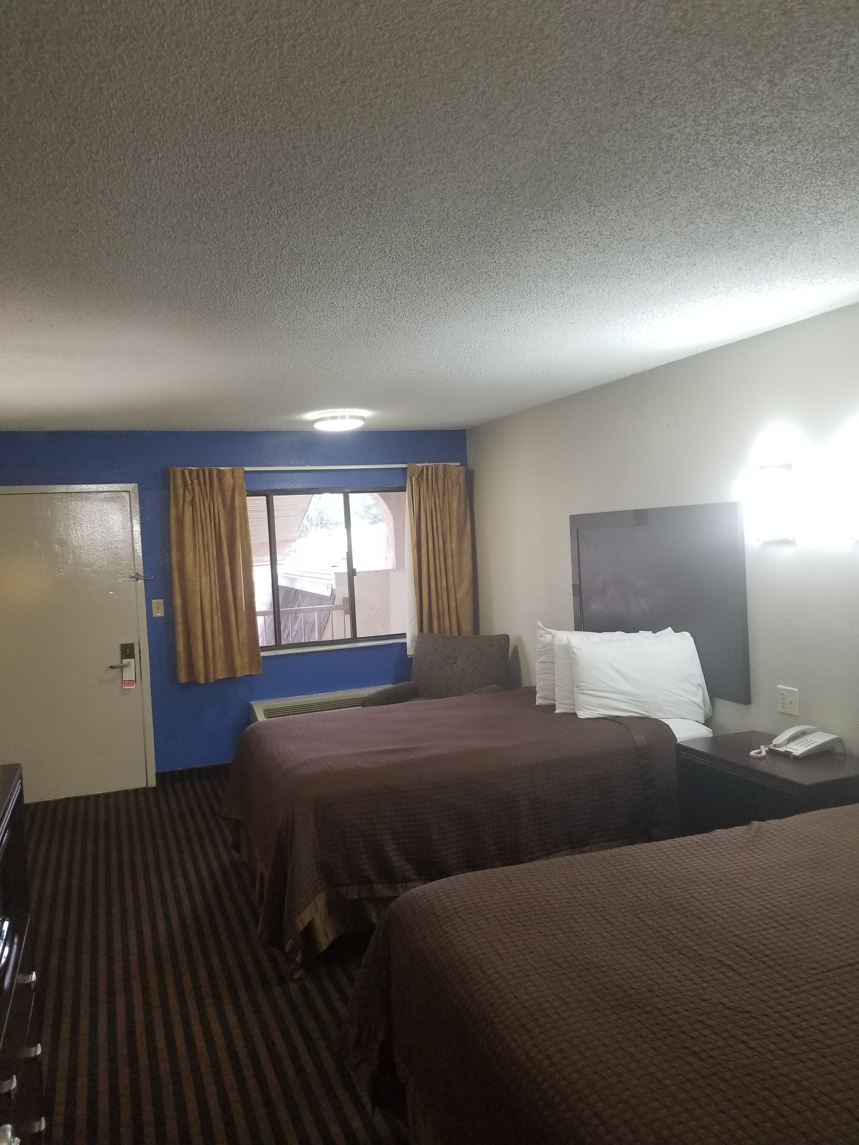 A hotel room with two beds, a window with curtains, a bedside table with a phone, and a door with a sign.