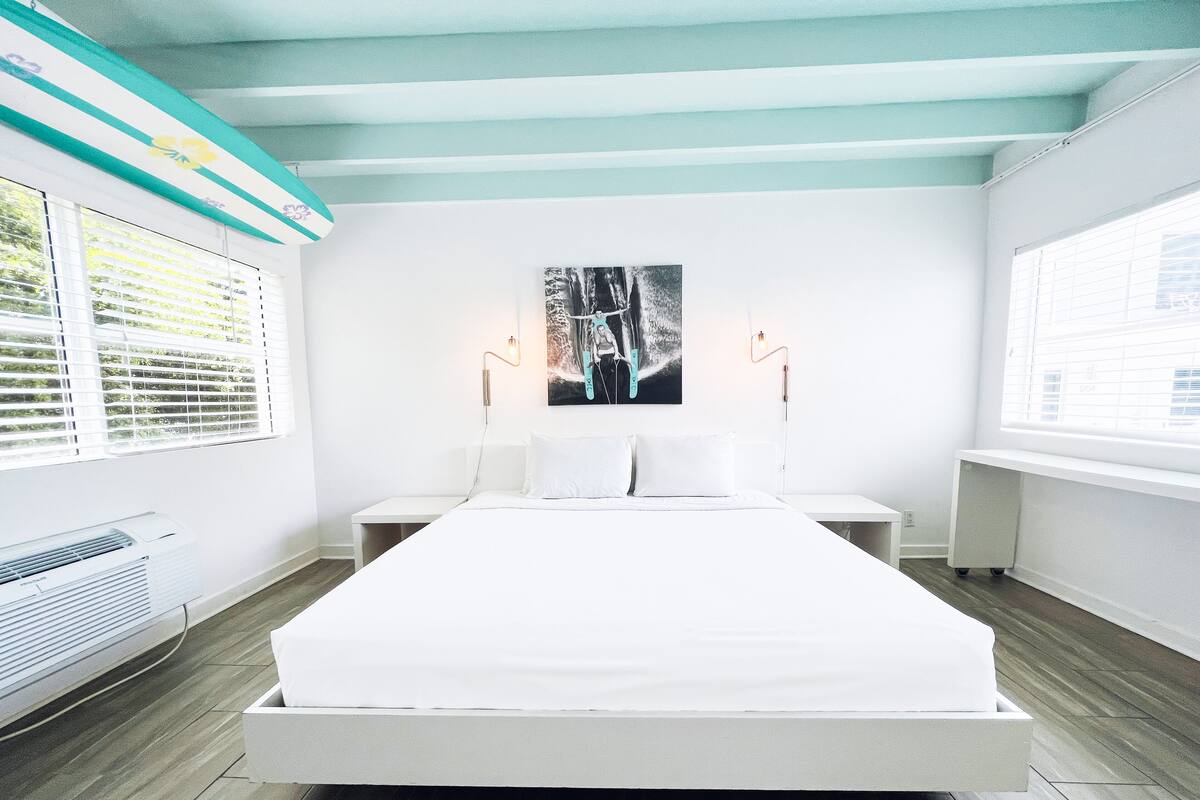 Une chambre à coucher moderne avec un grand lit, une œuvre d’art accrochée au mur et une fenêtre équipée de stores.