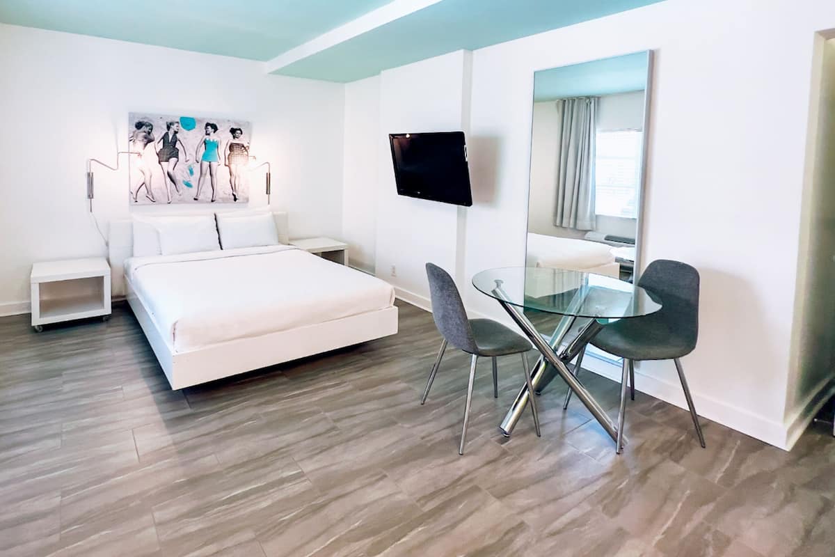 Une chambre d’hôtel moderne dotée d’un grand lit, d’une table à manger en verre avec deux chaises, d’un téléviseur à écran plat fixé au mur et d’un grand miroir.