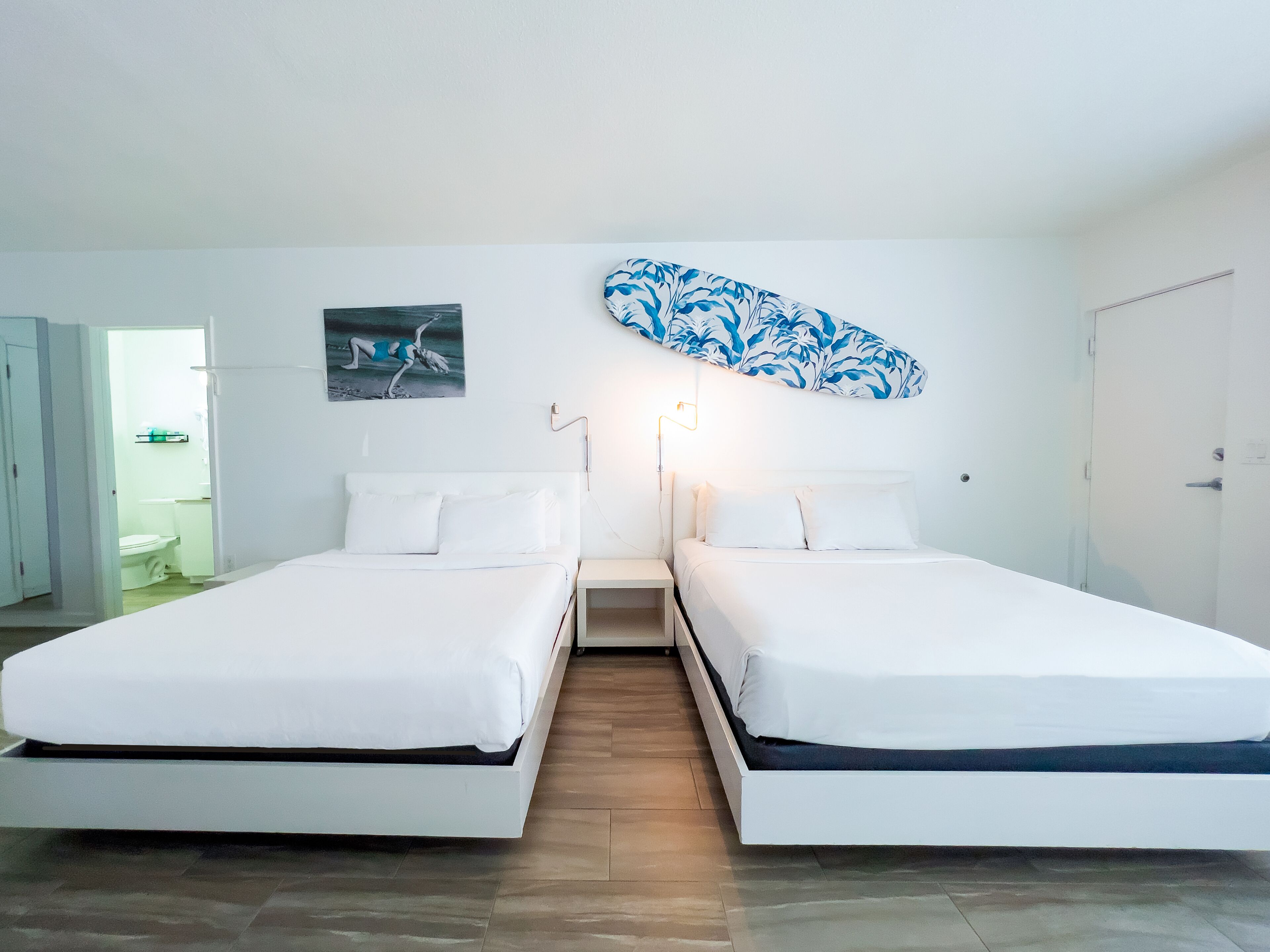 Habitación de hotel con dos camas individuales, un baño visible a través de una puerta abierta y una obra de arte colgada en la pared.