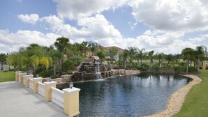 A landscaped pond with a waterfall, surrounded by palm trees and a paved walkway. - Paradise Palms Resort by Global Resort Homes (Kissimmee)