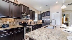 A modern kitchen with dark cabinets, granite countertops, stainless steel appliances, and a large island with a sink. - Paradise Palms Resort by Global Resort Homes (Kissimmee)