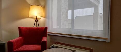 Una habitación con un sillón rojo, una mesa, una lámpara de pie y una ventana con persianas.