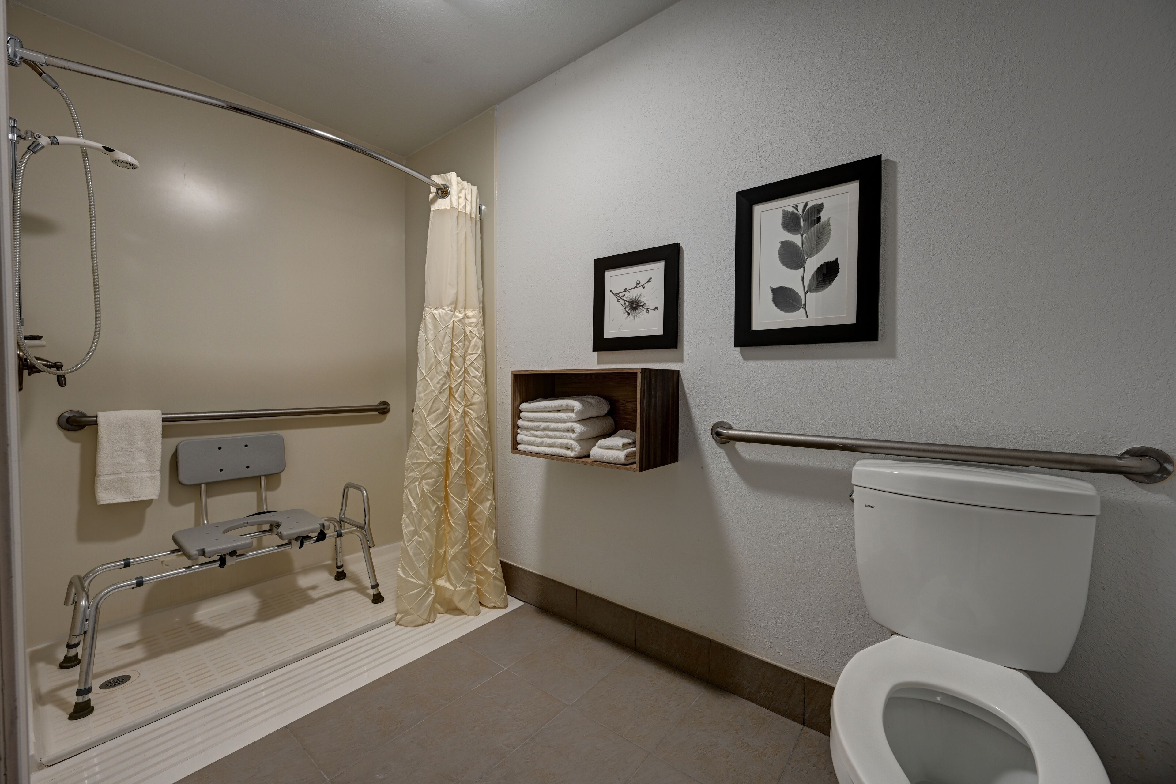 A bathroom with a shower, toilet, and a grab bar. There are towels and a shelf with folded towels.