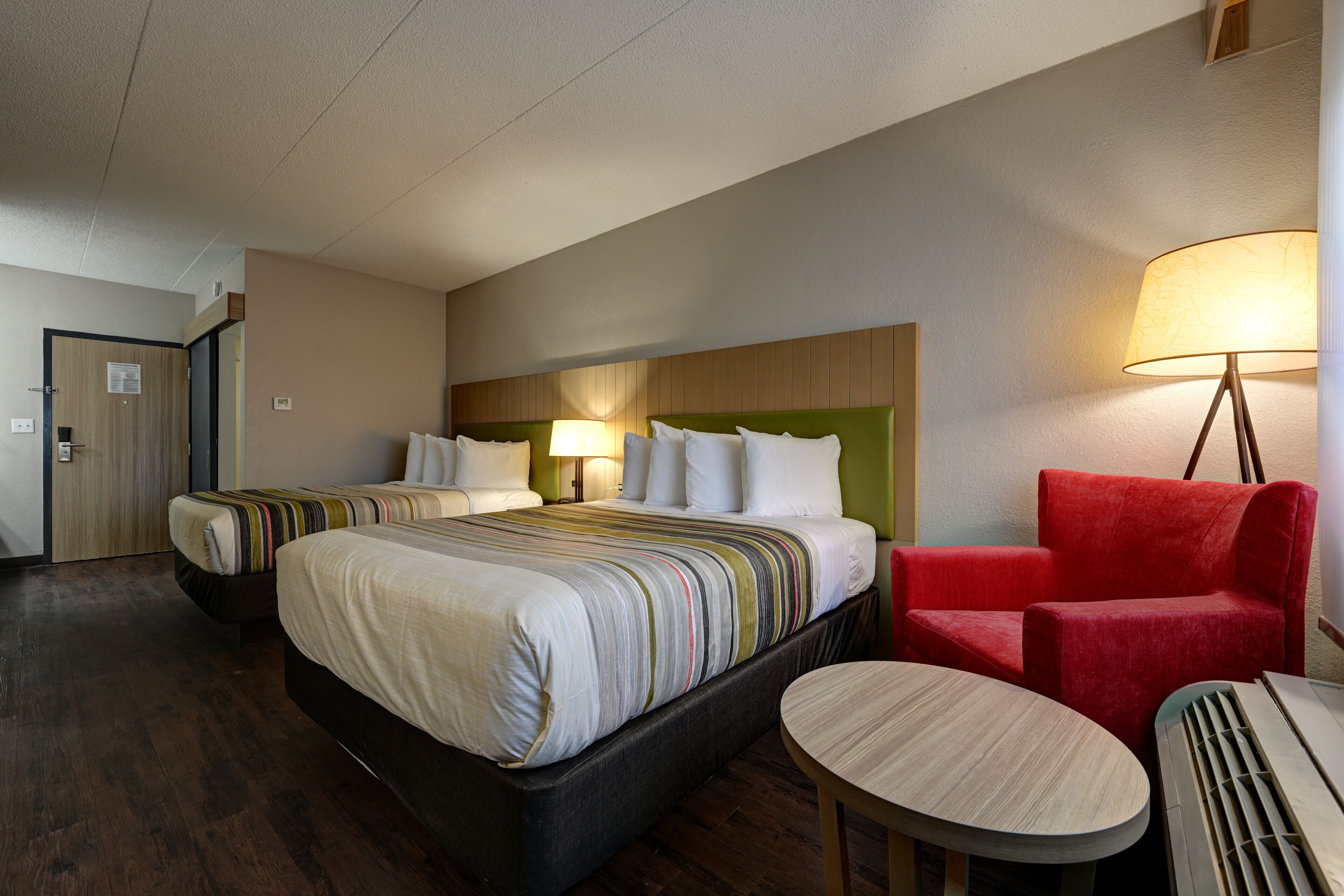 A hotel room with two beds, a red armchair, a small round table, and a lamp.