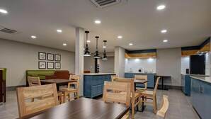 Un espace repas moderne avec des tables et des chaises en bois, un coin salon avec des banquettes vertes, et une cuisine dotée de meubles bleus et d’un réfrigérateur noir.