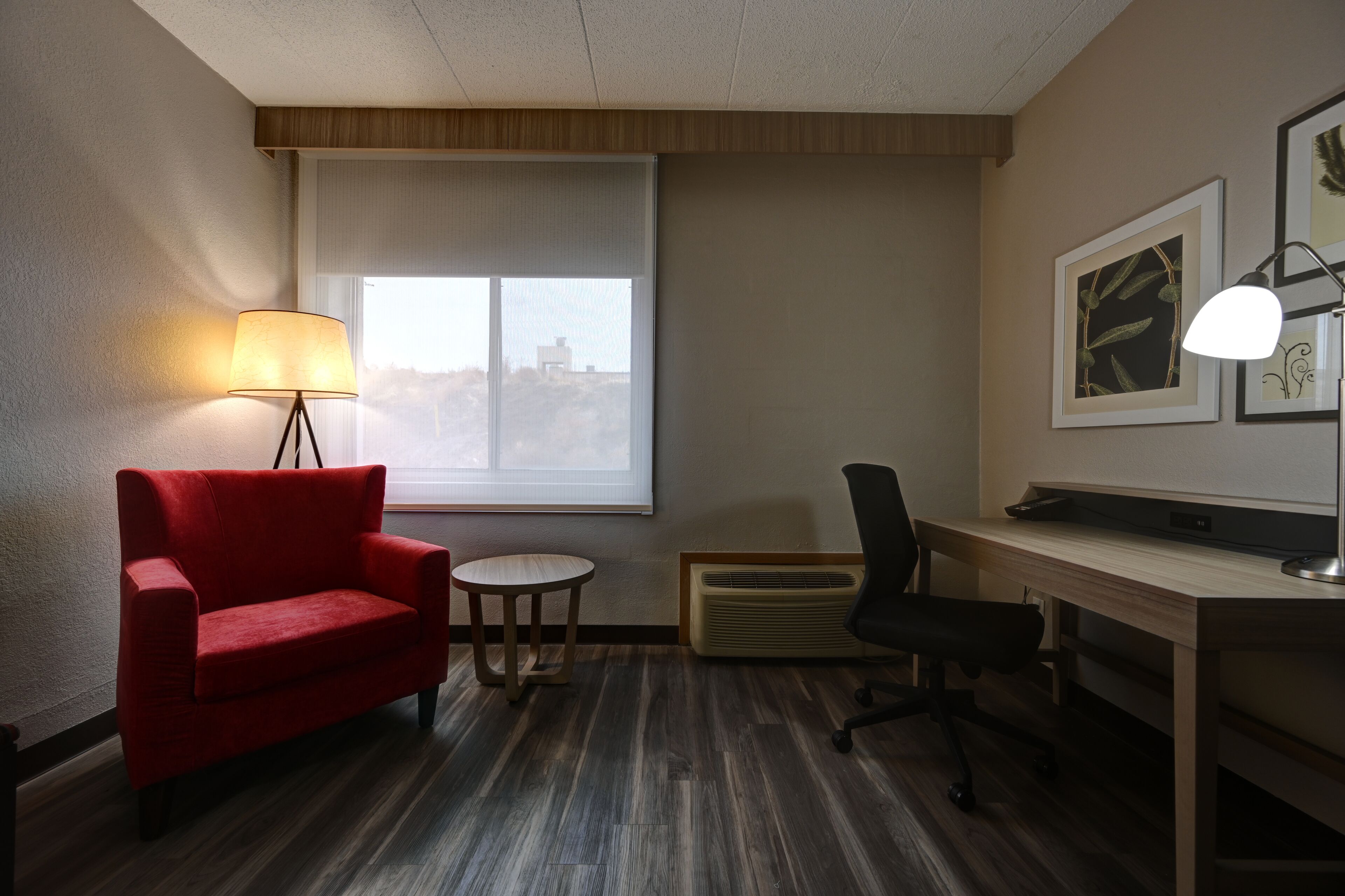 Habitación de hotel con sofá rojo, una pequeña mesa redonda, escritorio con computadora, lámpara de pie y un amplio ventanal con persianas.
