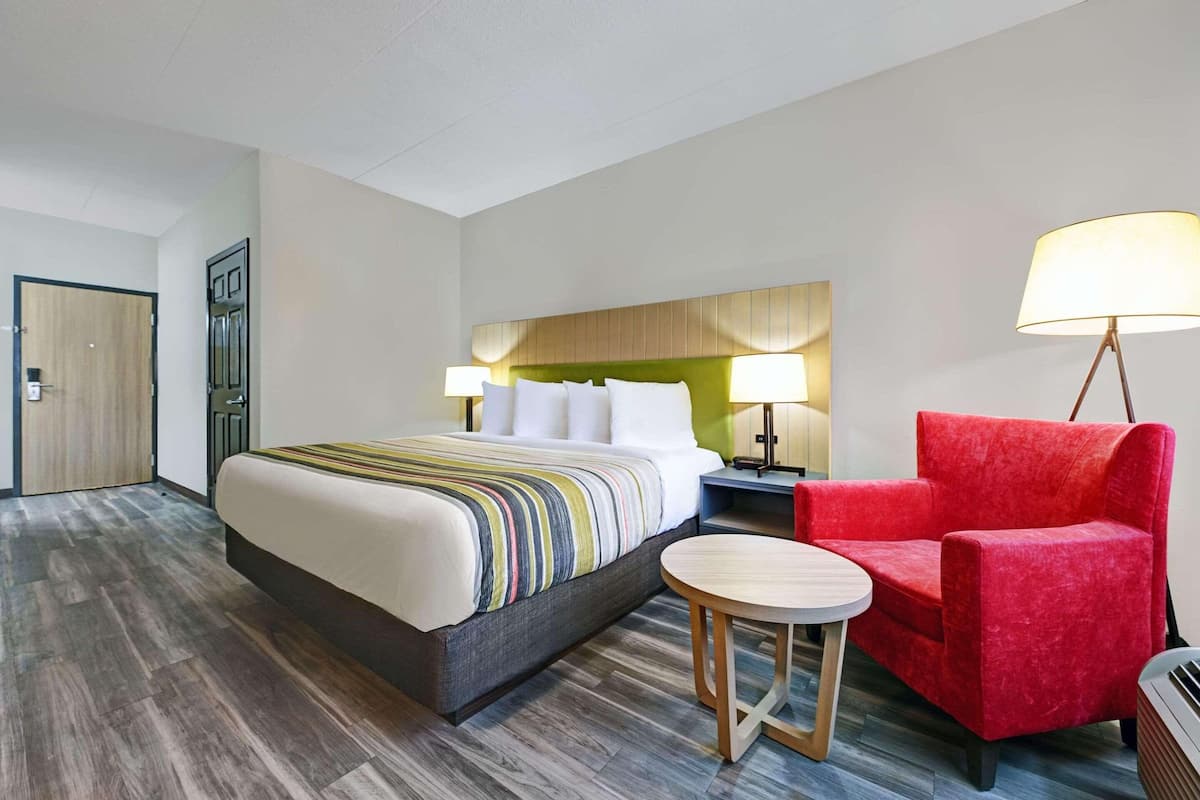 A hotel room with a bed, bedside table, lamp, and a red chair.