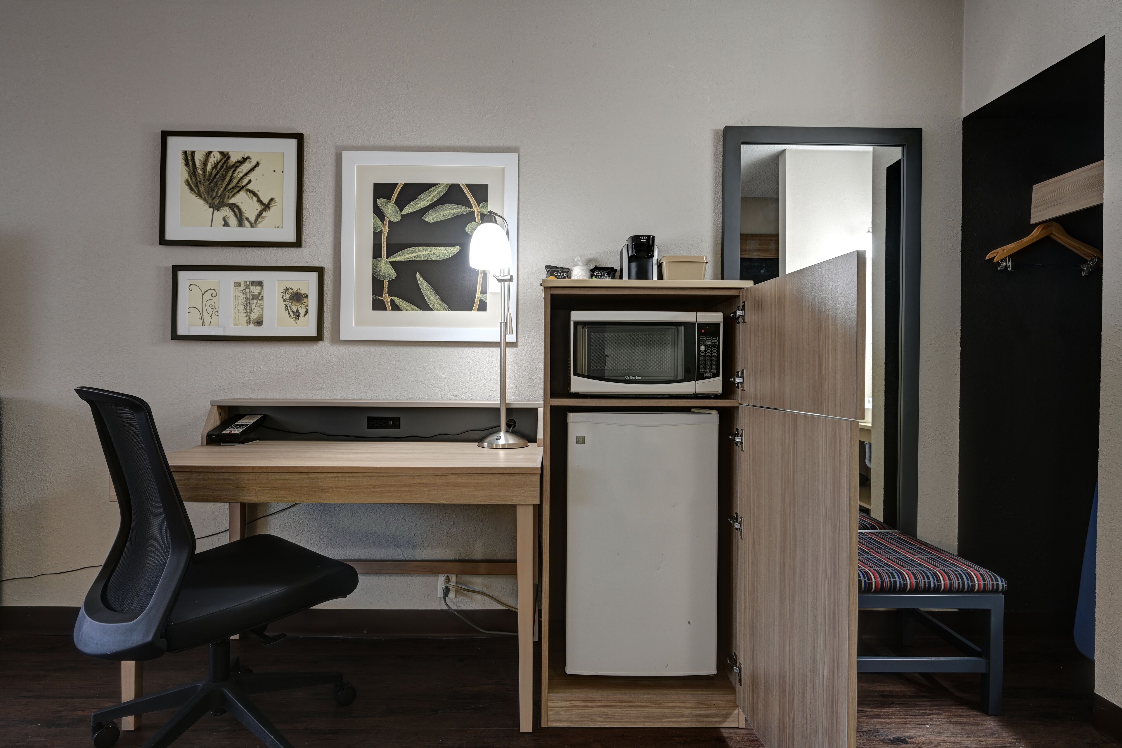 Una habitación compacta con un escritorio de madera, una silla de oficina negra, un refrigerador mini blanco, un microondas y una cama pequeña con un cojín a rayas.