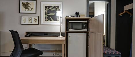 Una habitación compacta con un escritorio de madera, una silla de oficina negra, un refrigerador mini blanco, un microondas y una cama pequeña con un cojín a rayas.