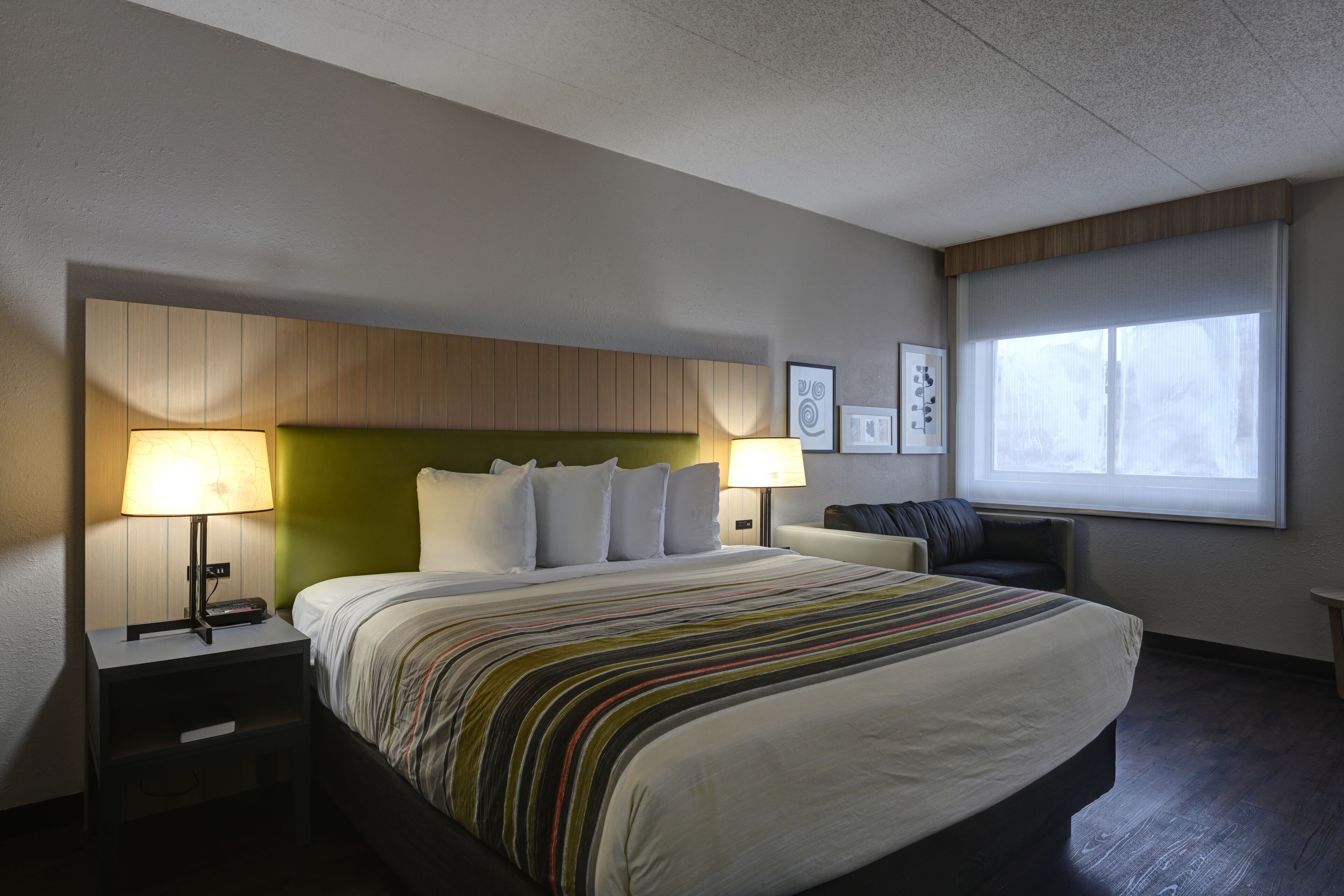 A hotel room with a large bed, two bedside tables with lamps, a green headboard, a window with a view, and a small seating area with a chair and a table.