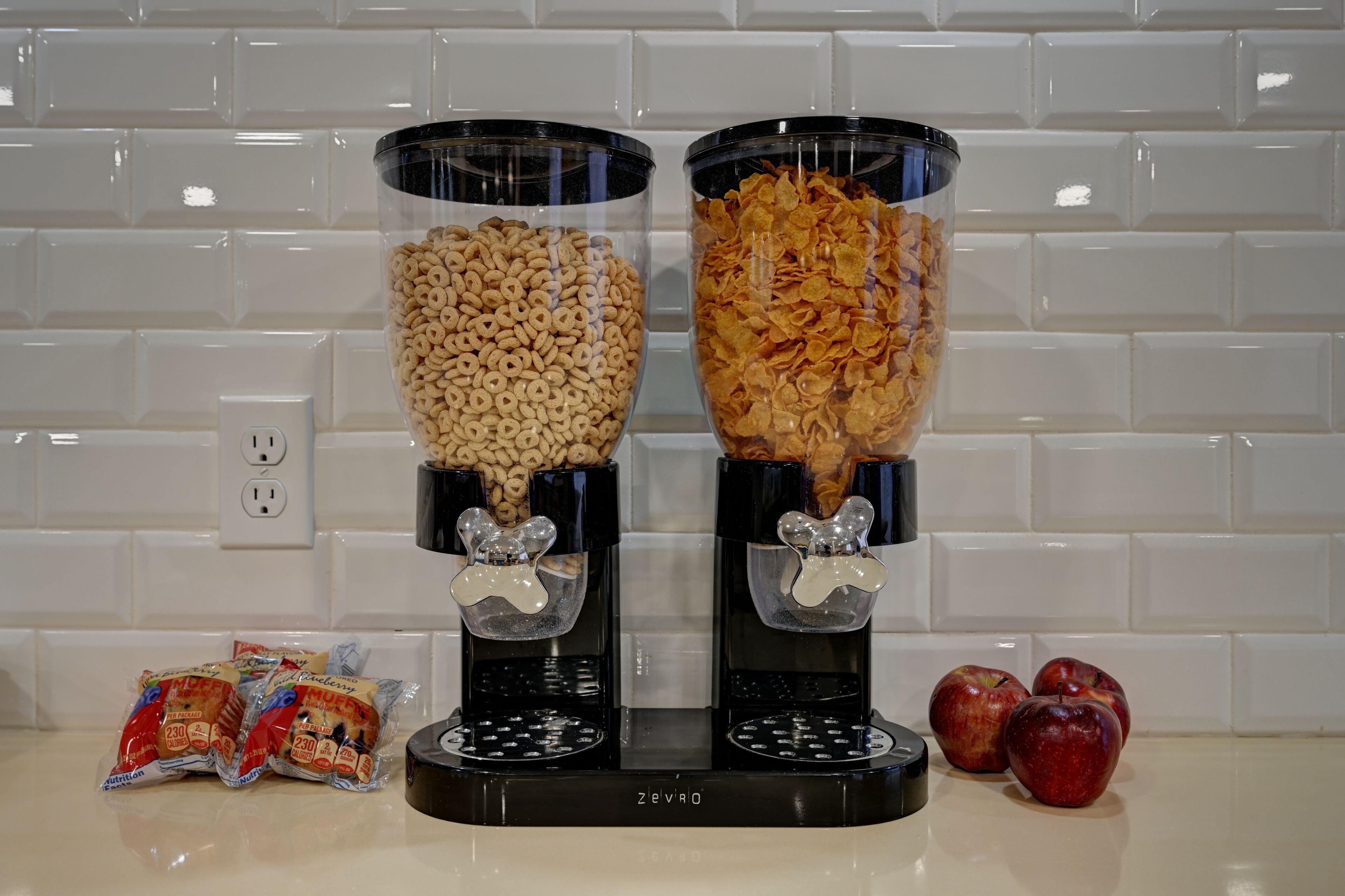 Two cereal dispensers with a white tiled backsplash and a power outlet in the background.
