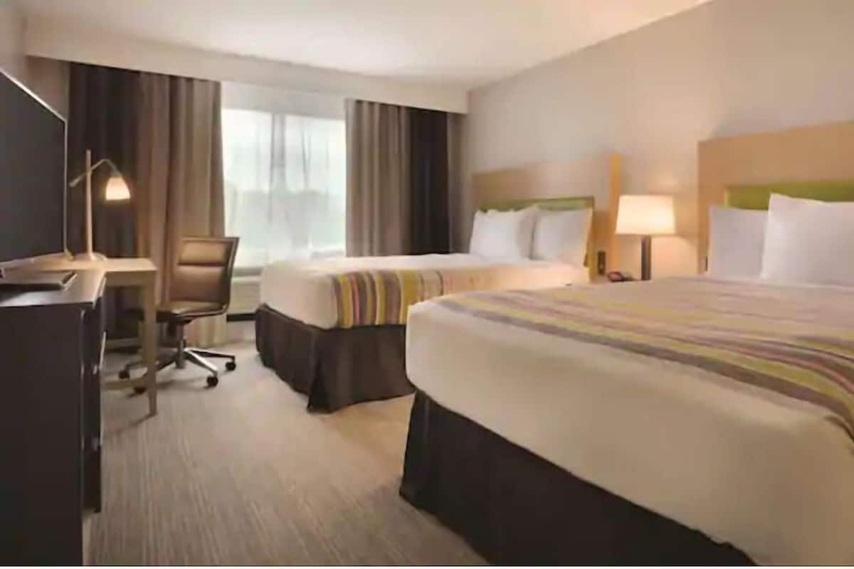 A hotel room with two beds, a desk, a chair, and a lamp.