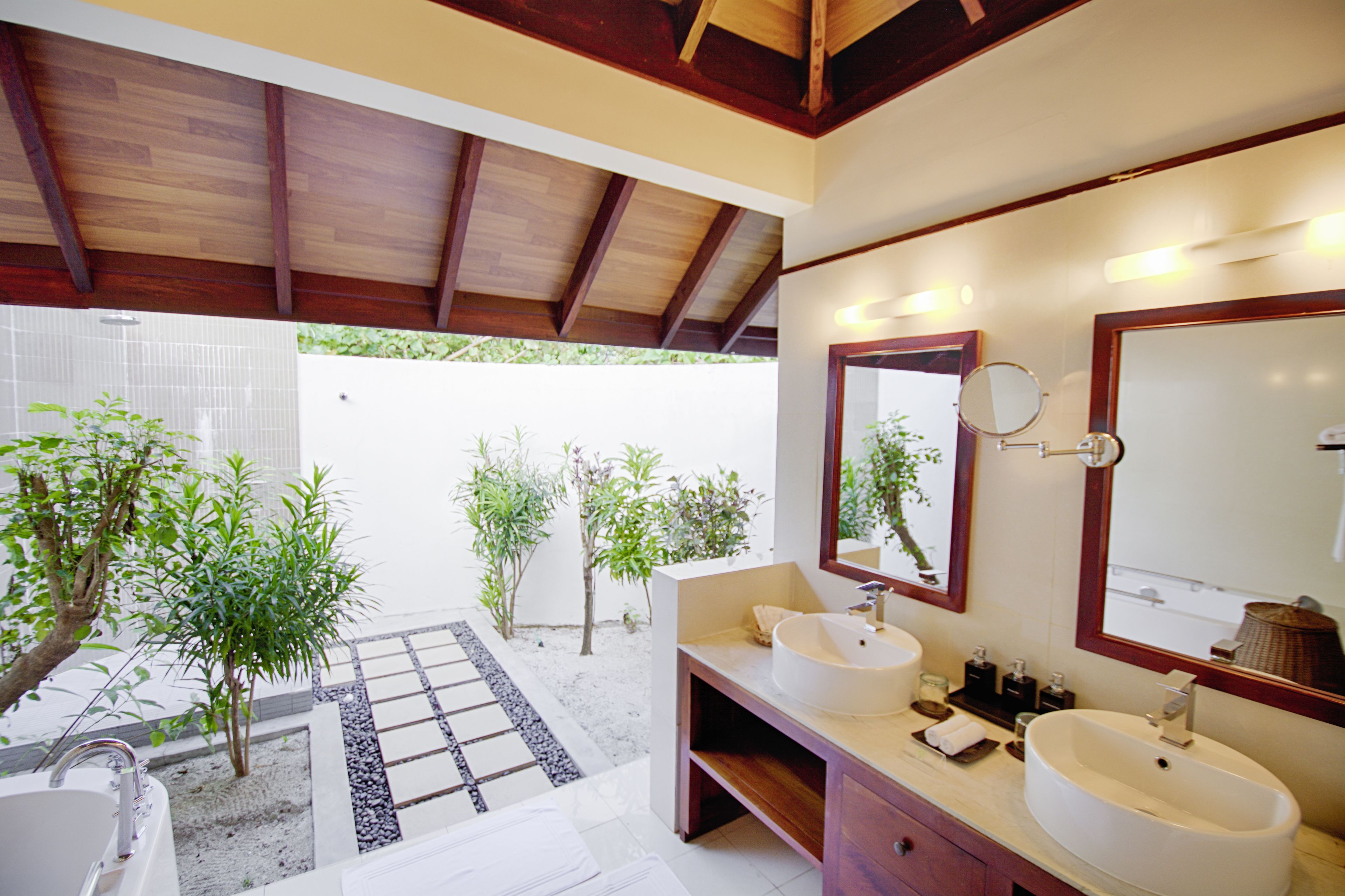 A modern bathroom with two sinks, a large mirror, and a view of a garden with small trees.