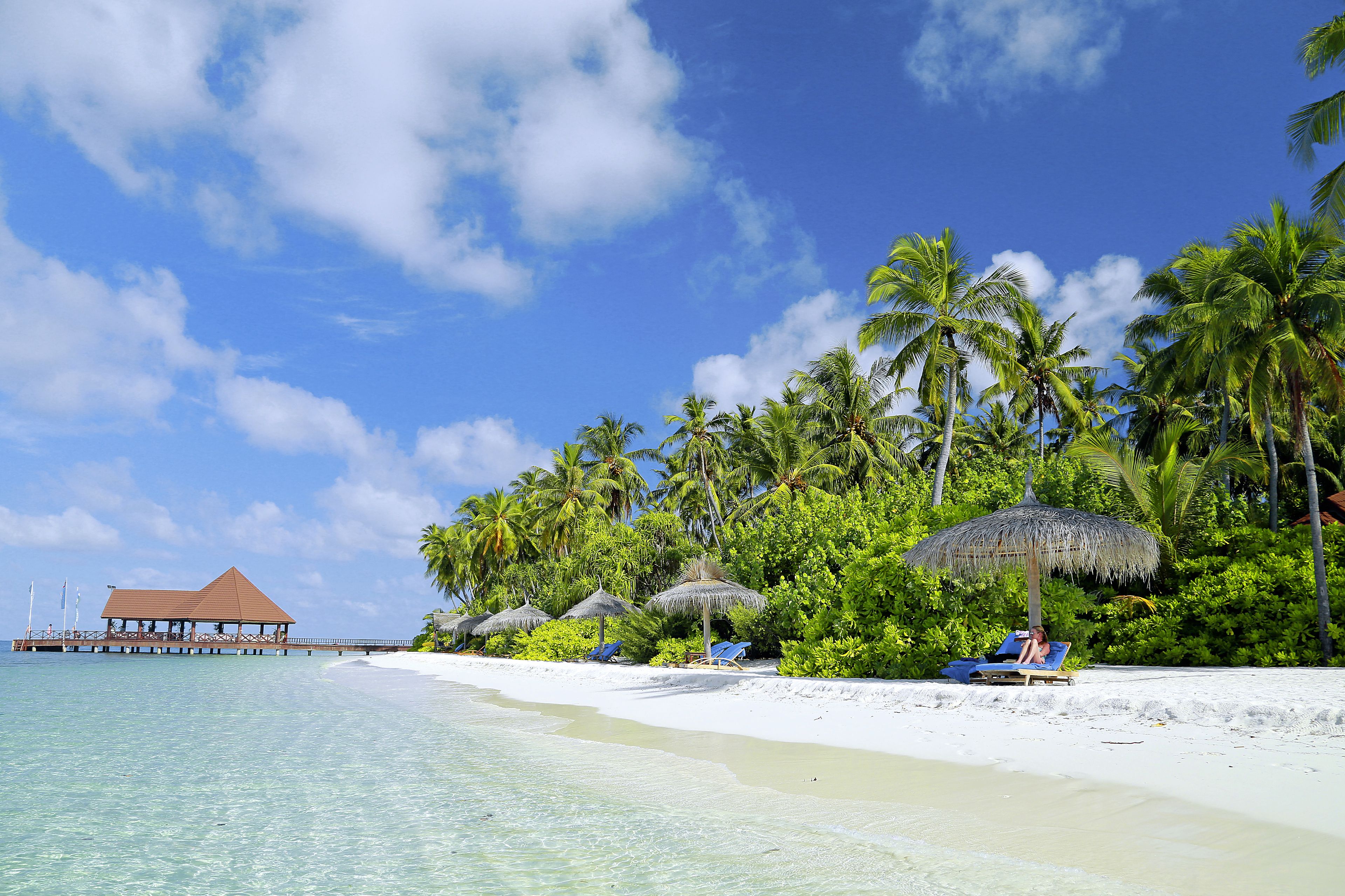 Een strand met helder turquoise water, wit zand en palmbomen. Er staan rieten hutten en ligbedden langs de kust.