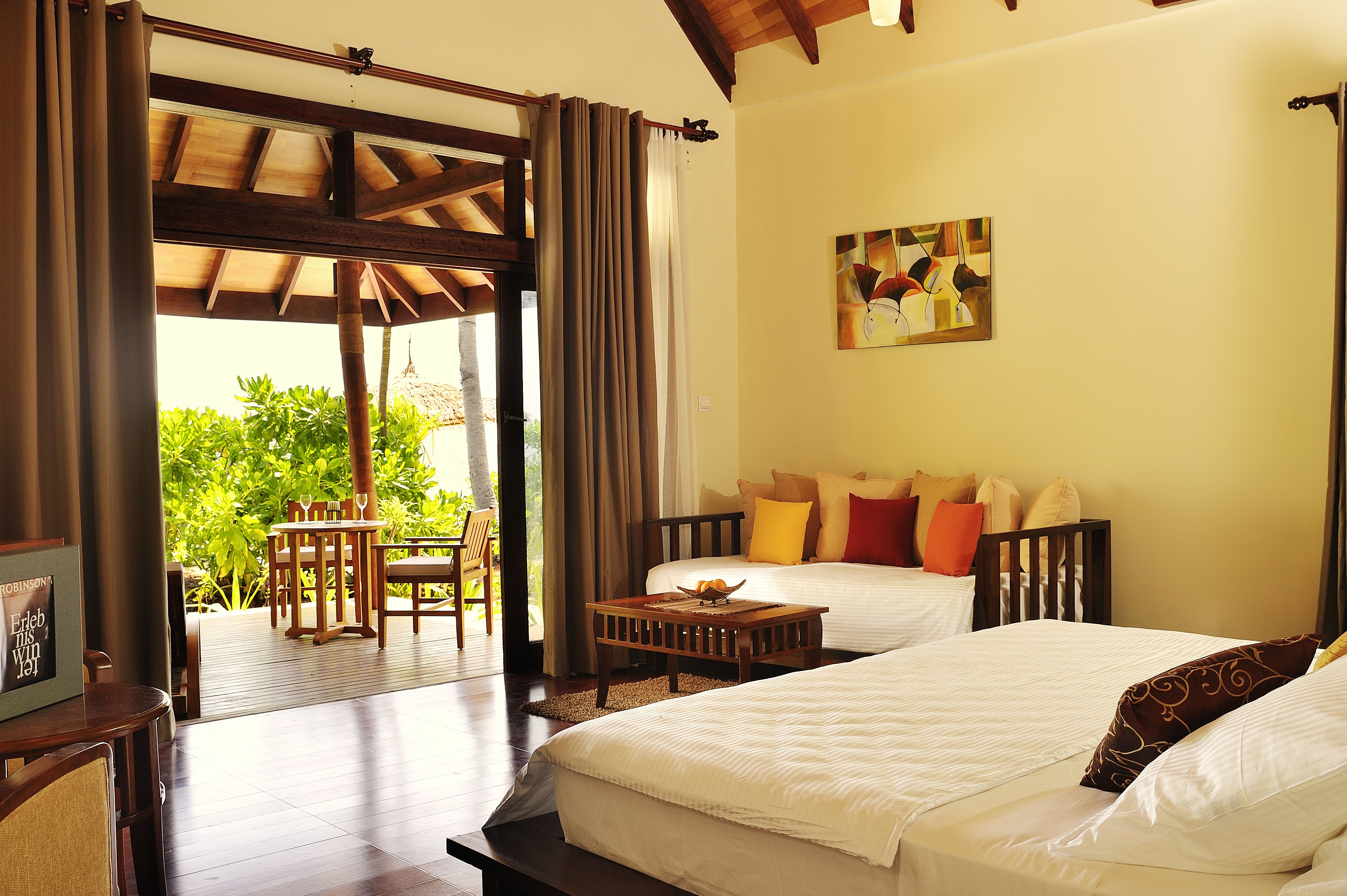 A hotel room with two beds, a wooden ceiling, a dining area with a table and chairs, and a view of greenery outside.