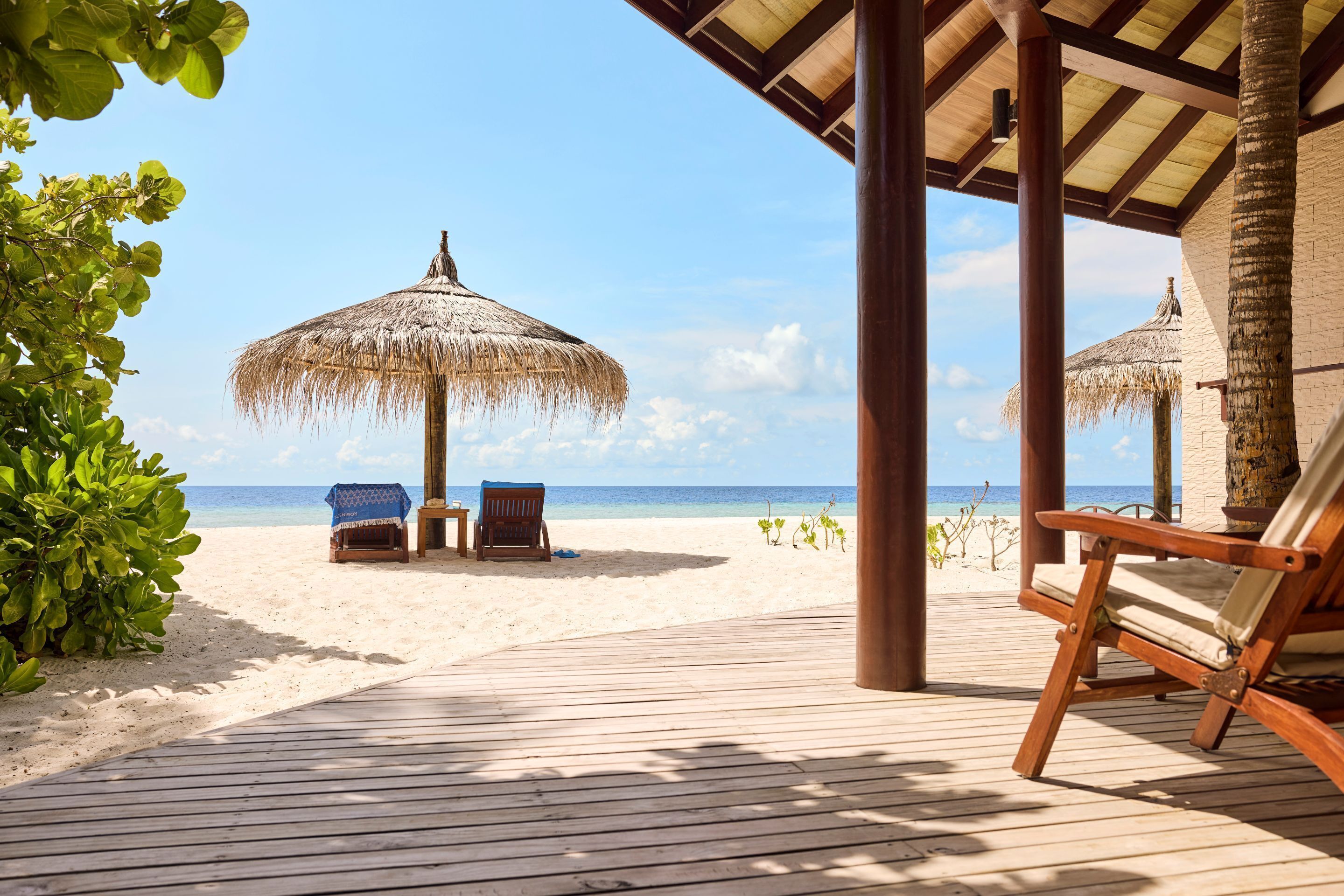 A wooden deck with two lounge chairs and a thatched umbrella, overlooking a sandy beach and clear blue sea.