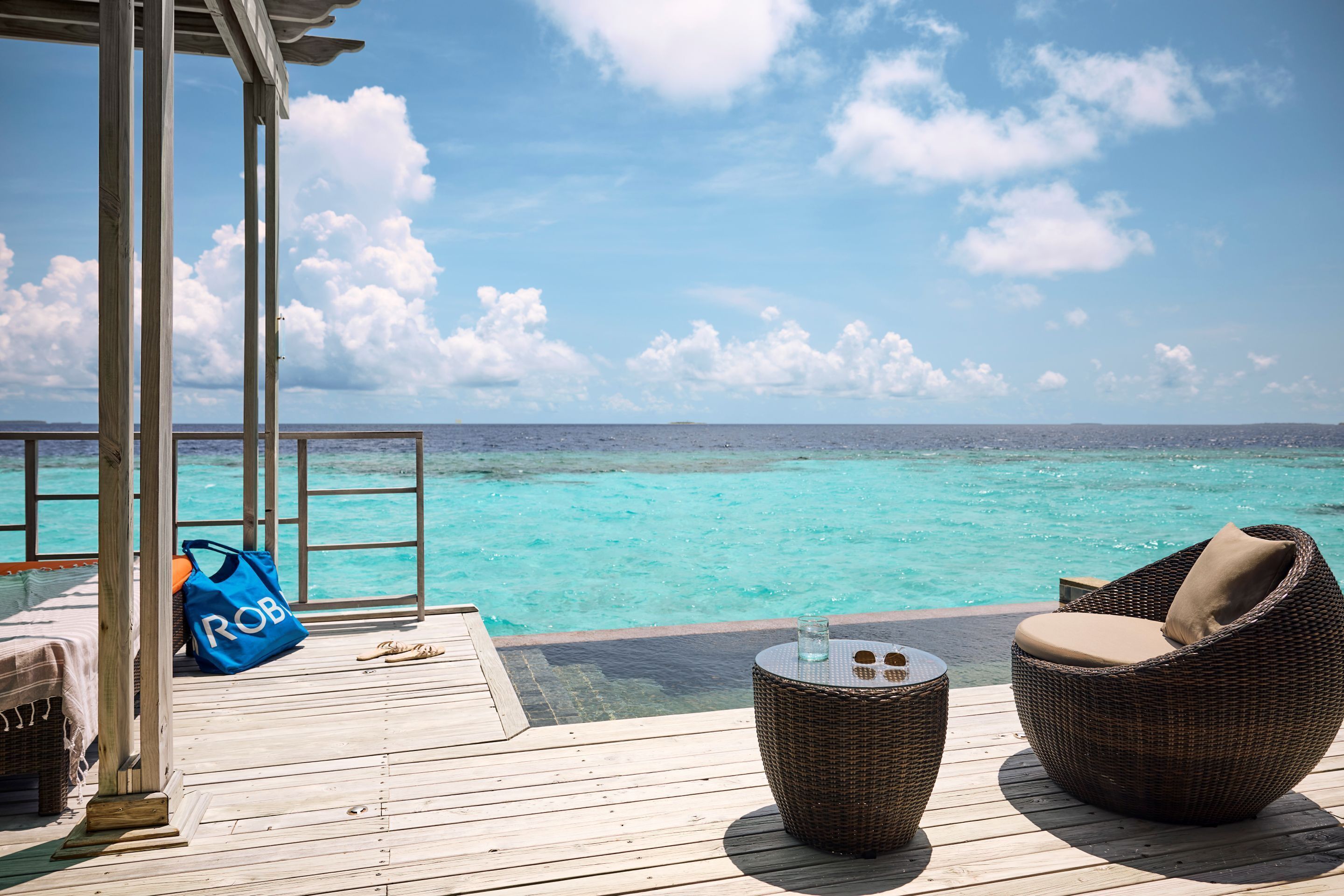 A wooden deck with a wicker chair and a small table, overlooking a clear turquoise sea.