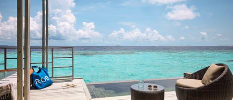 A wooden deck with a wicker chair and a small table, overlooking a clear turquoise sea.