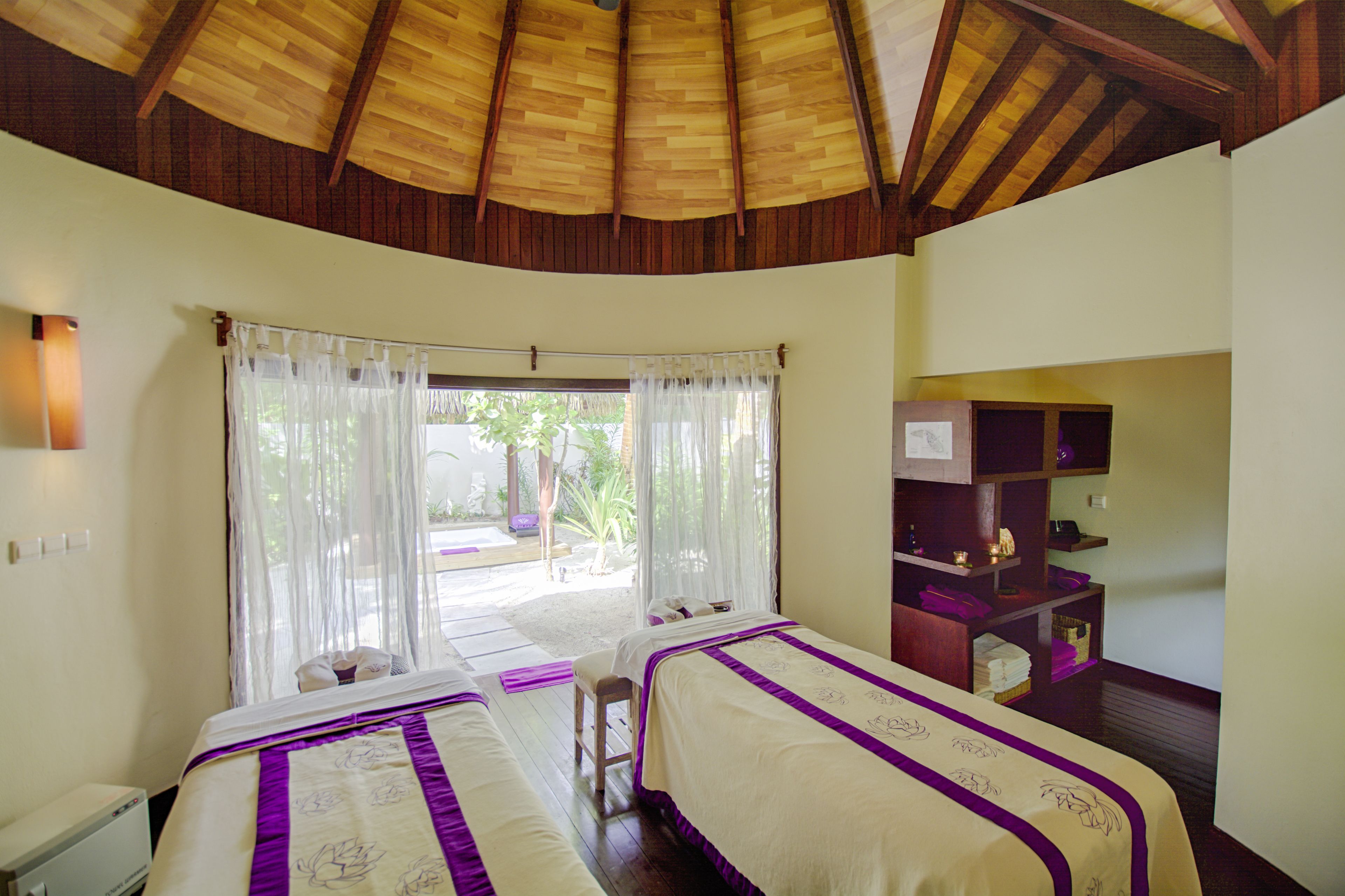 A room with two beds, a wooden ceiling, a window with sheer curtains, and a small table with a lamp.
