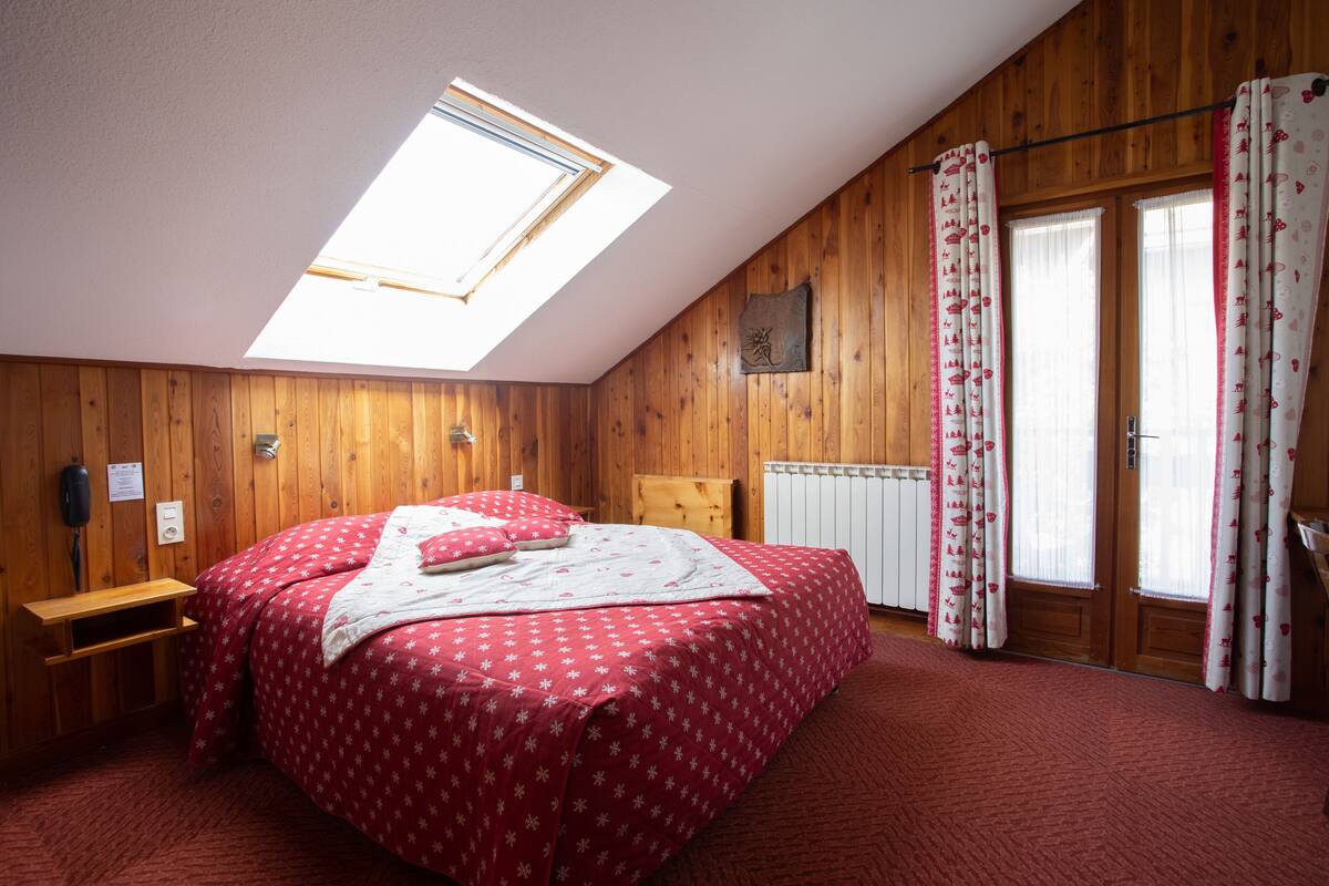 A bedroom with a skylight, wooden walls, a bed with red bedding, a wooden bench, and a window with curtains.