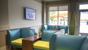 Un hall de hotel con asientos azules y amarillos, un televisor de pantalla plana en la pared y una ventana con un paraguas afuera.