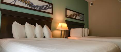 A hotel room with two beds, a headboard with a decorative panel, a wall-mounted lamp, and two framed pictures of a roller coaster.