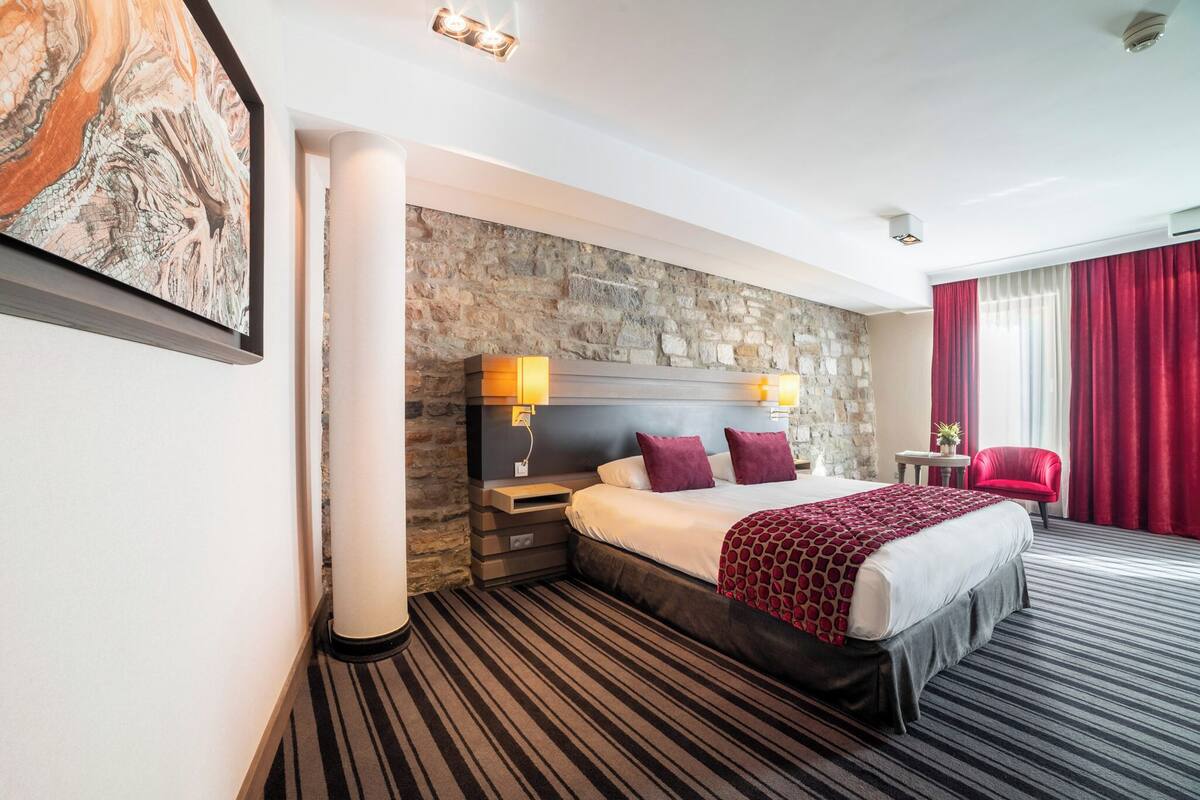 Habitación de hotel con pared de piedra, una cama grande con manta roja, una silla roja y una mesita con una planta.