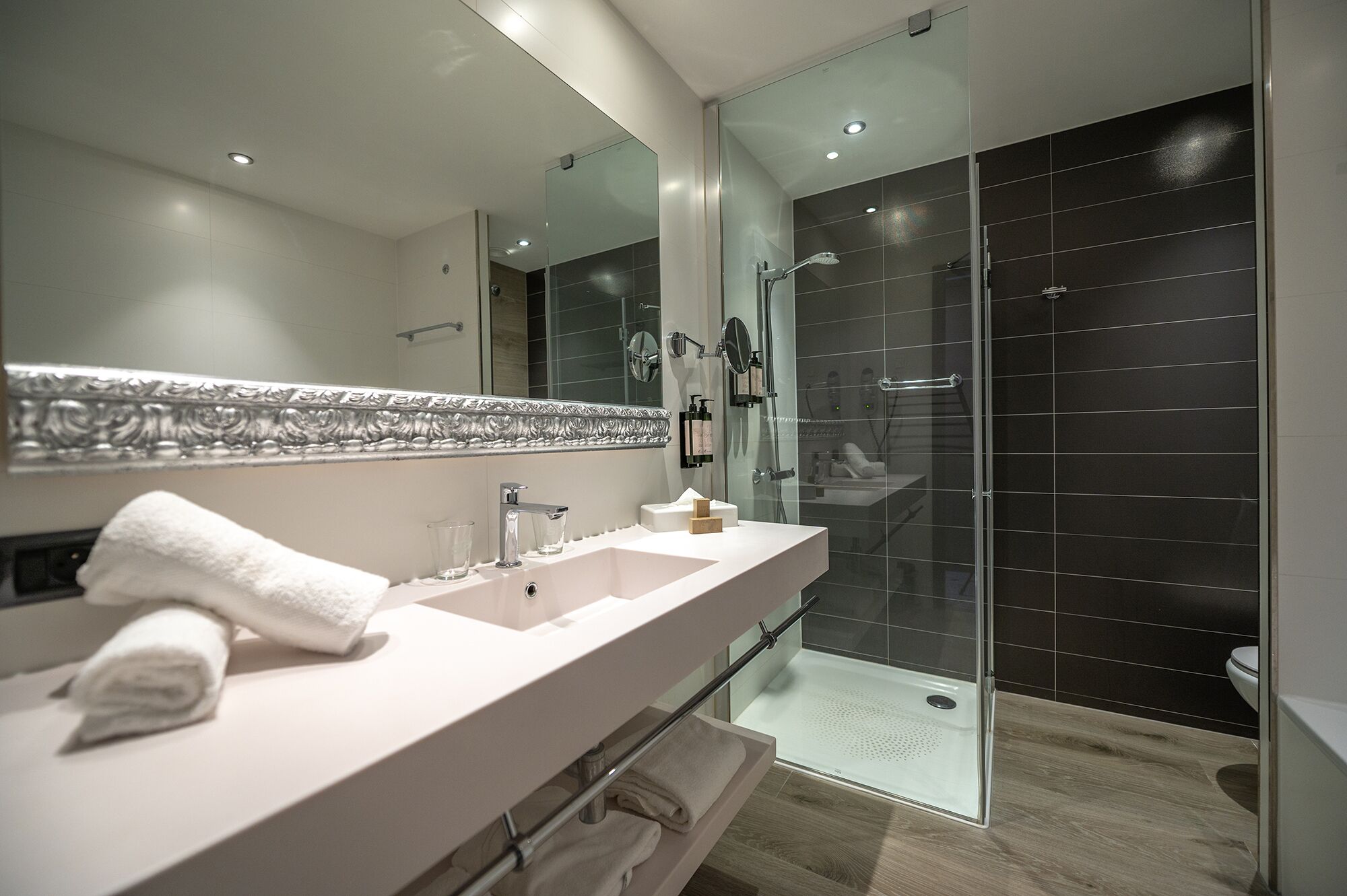 Een moderne badkamer met een grote spiegel, een witte wastafel, een douchecabine van glas en een houten vloer.
