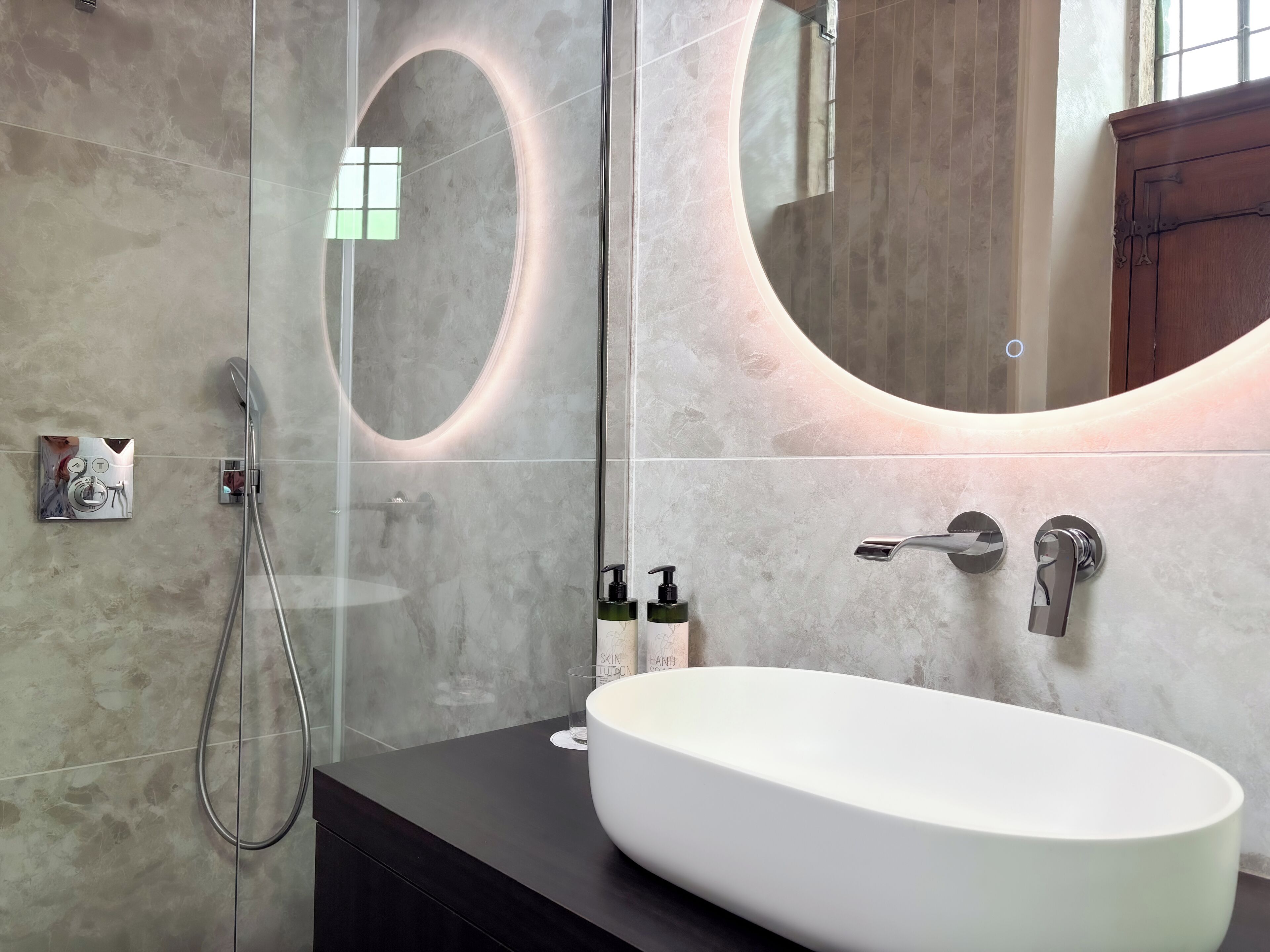 Un baño moderno con un lavamanos ovalado grande, un cabezal de ducha de mano y un espejo montado en la pared con luz circular.