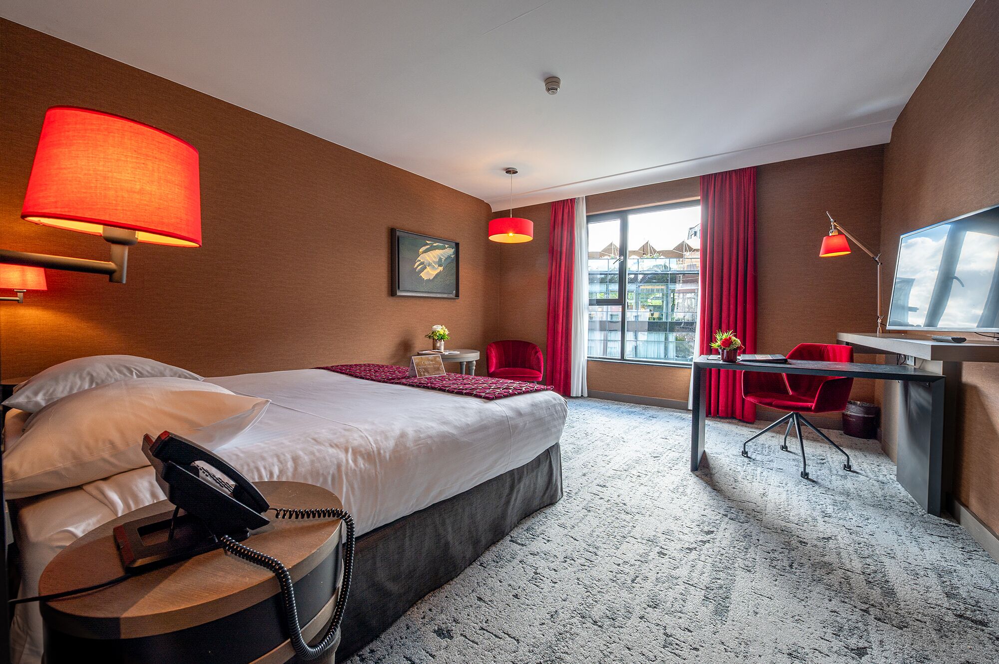 Une chambre d’hôtel avec un grand lit, un bureau avec une chaise, une lampe de bureau, un téléphone et une fenêtre avec des rideaux.