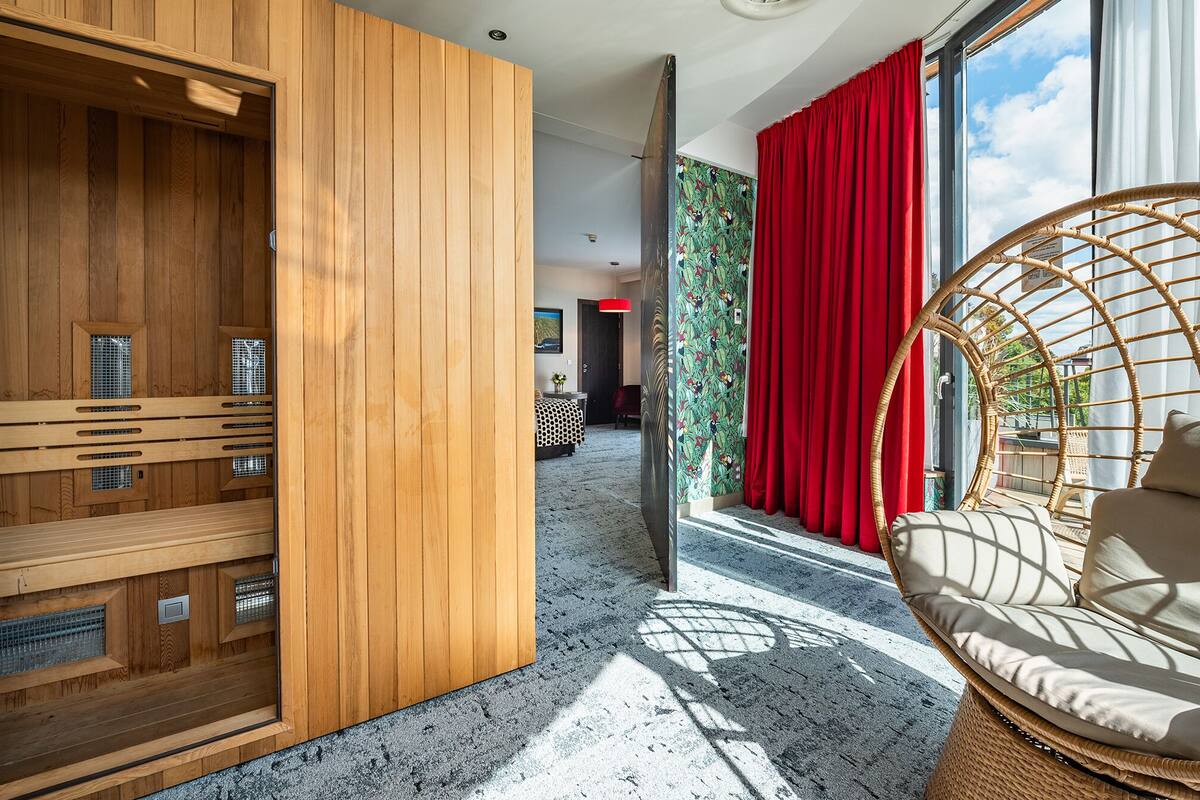 Habitación de hotel moderna con sauna de madera, cortina roja, silla de mimbre y vistas al exterior.
