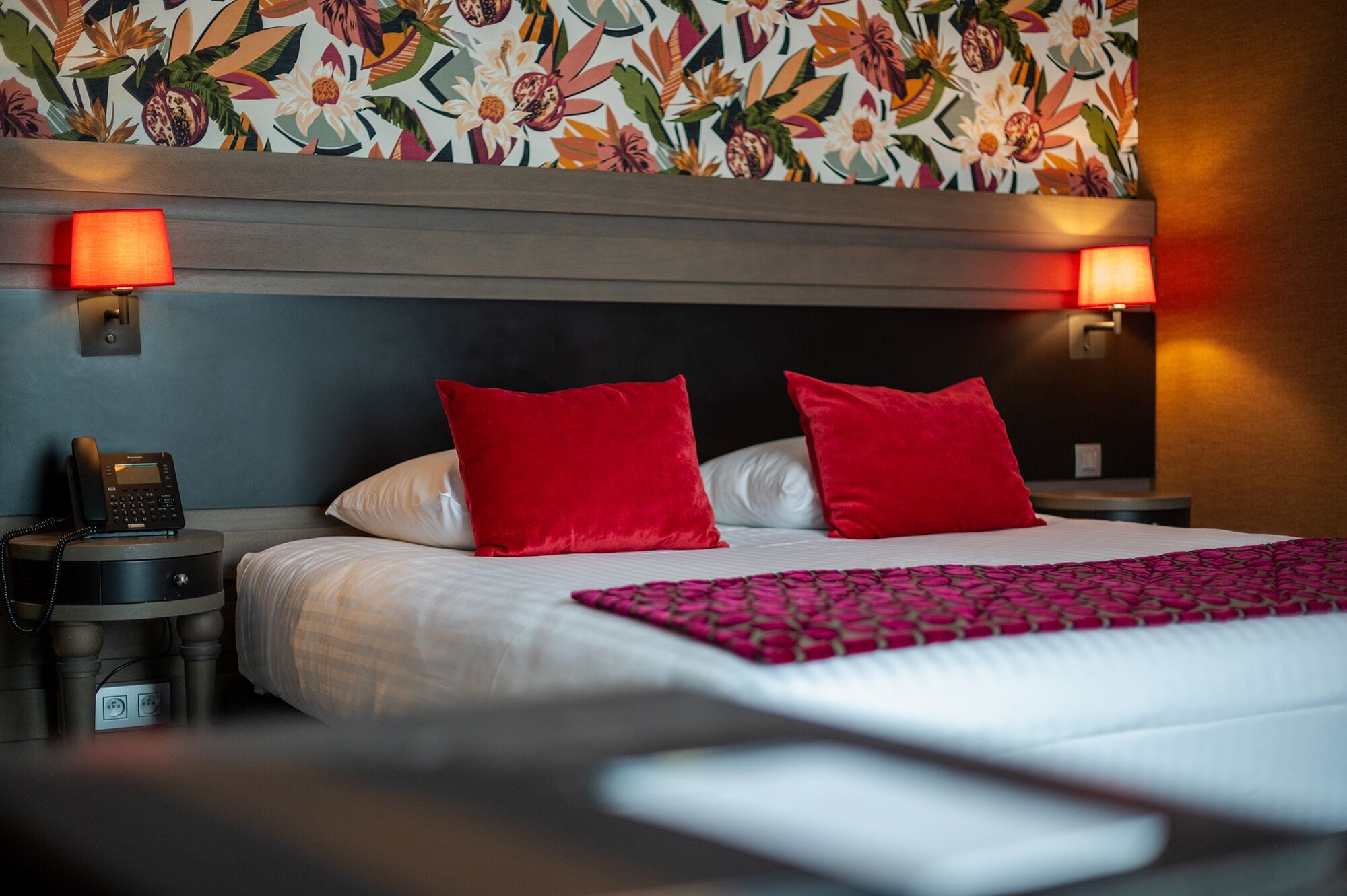 Habitación de hotel con una cama grande, dos almohadas rojas, un cabecero estampado, un teléfono en la mesita de noche y dos lámparas fijadas a la pared.