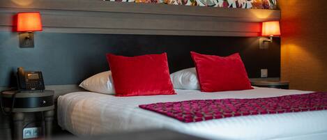 Habitación de hotel con una cama grande, dos almohadas rojas, un cabecero estampado, un teléfono en la mesita de noche y dos lámparas fijadas a la pared.